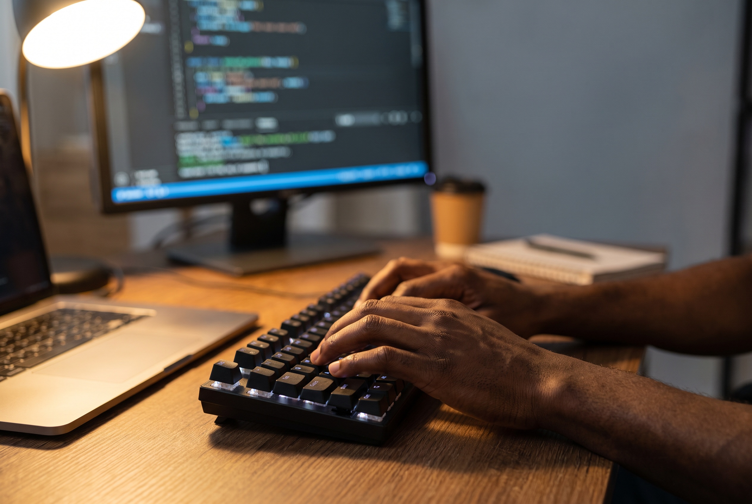 Person typing on a keyboard at a desk with a monitor displaying code, a laptop, and a cup in the background.