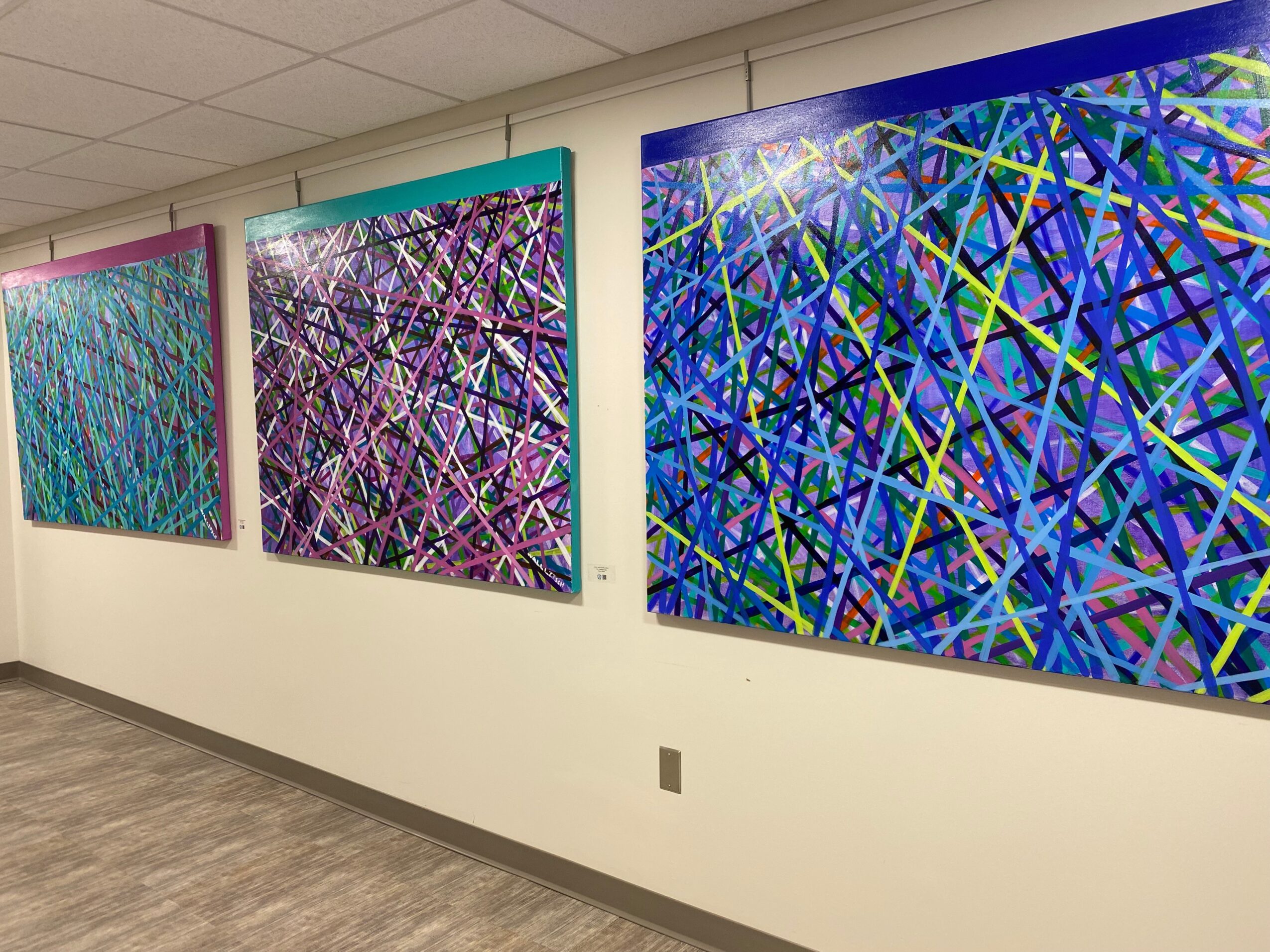 Three colorful abstract paintings with intersecting lines hang on a white wall in an art gallery.