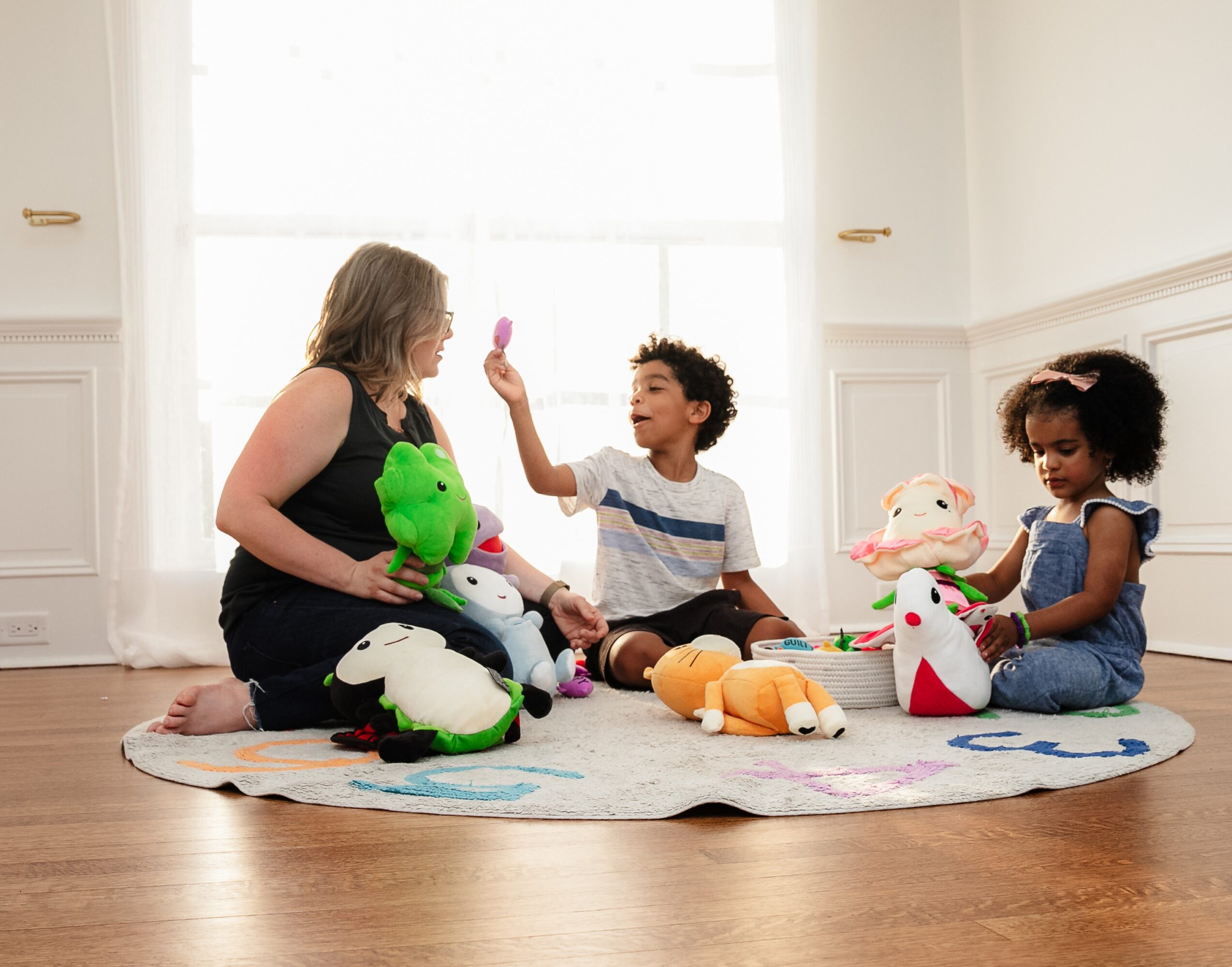 Woman and two children playing with plush toys on a round rug in a bright room.