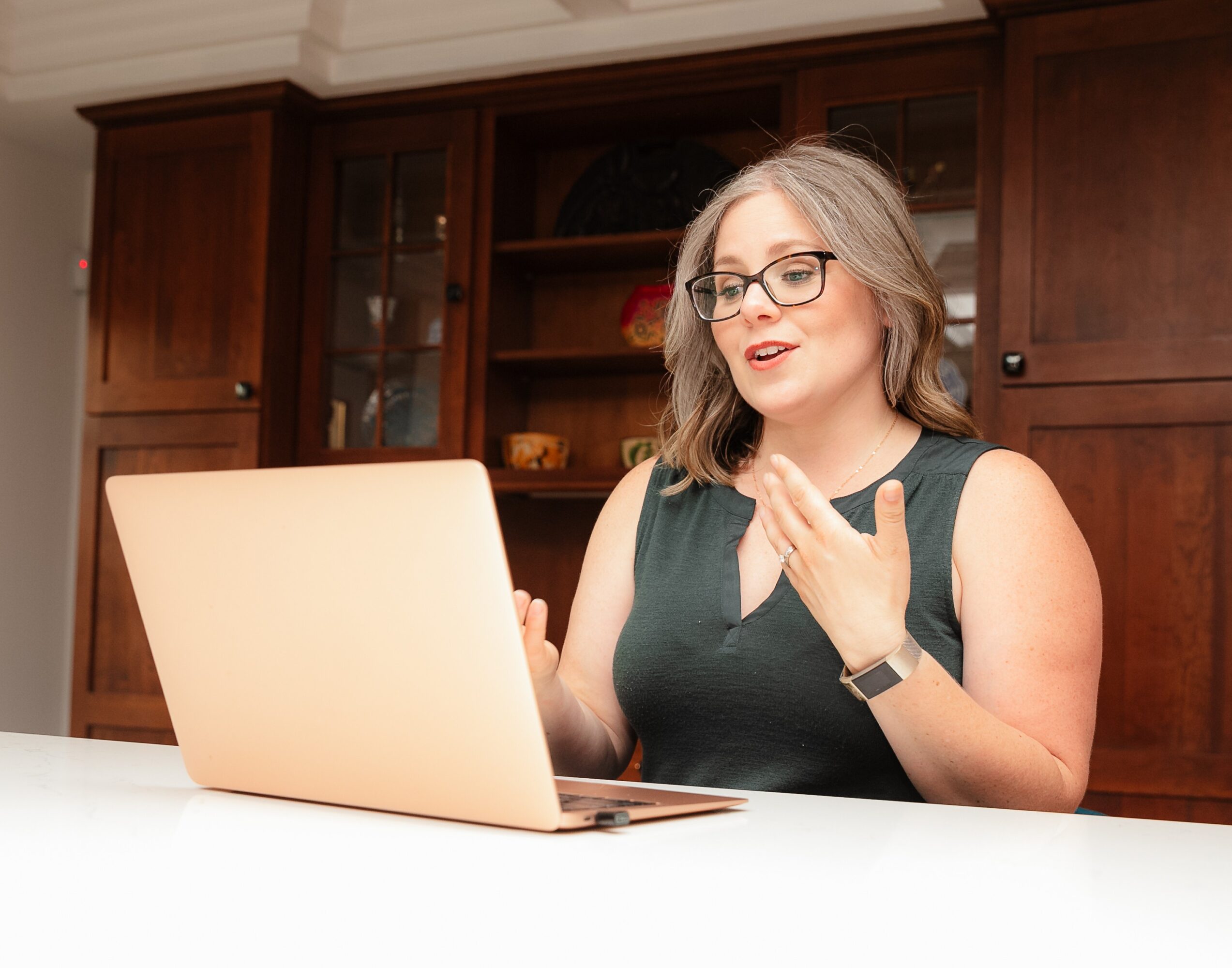 Woman with glasses and gray hair gestures while talking in front of a laptop, wooden cabinet background.