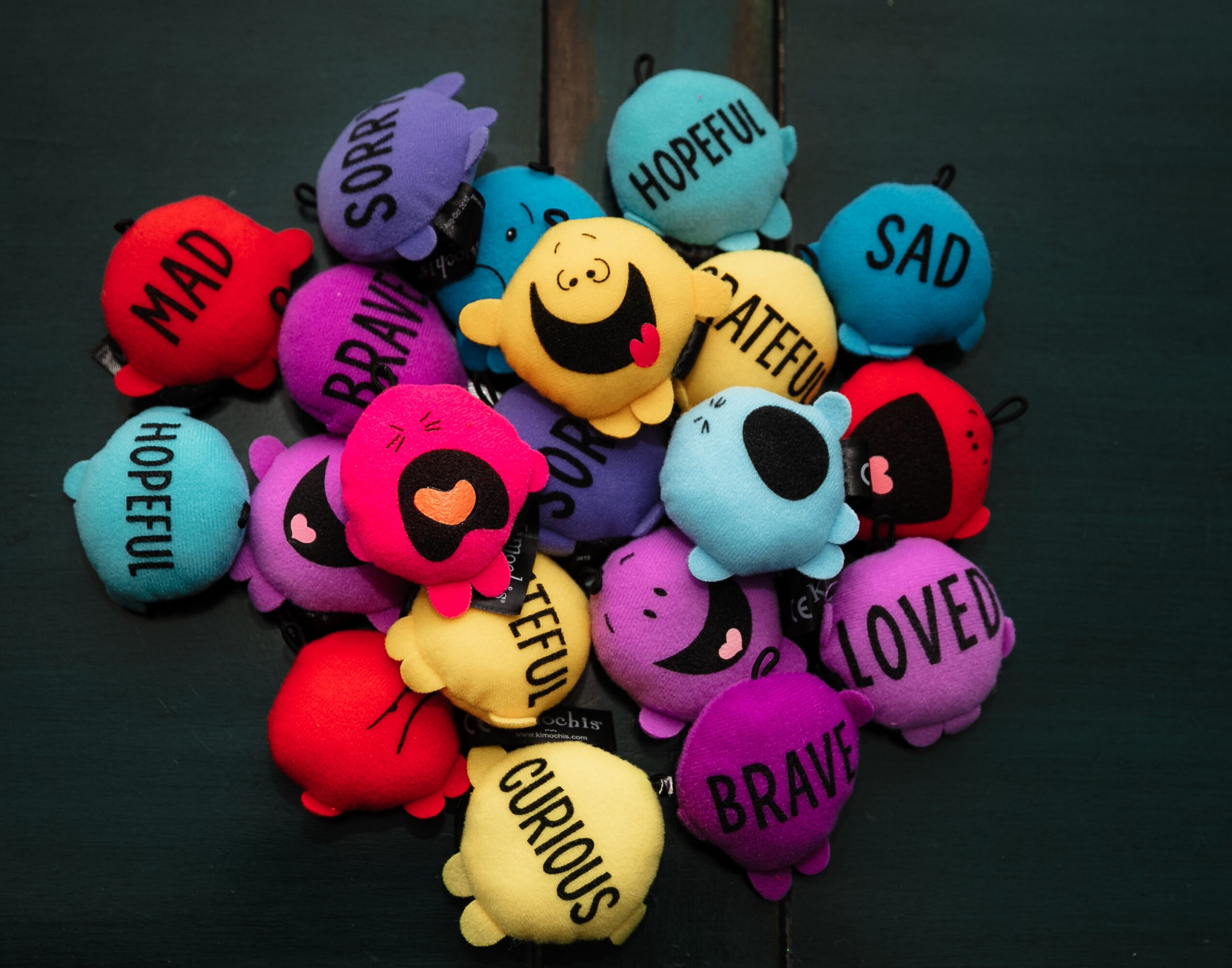 Colorful plush toys with words like 'happy', 'sad', 'brave', and 'curious' on a dark surface.