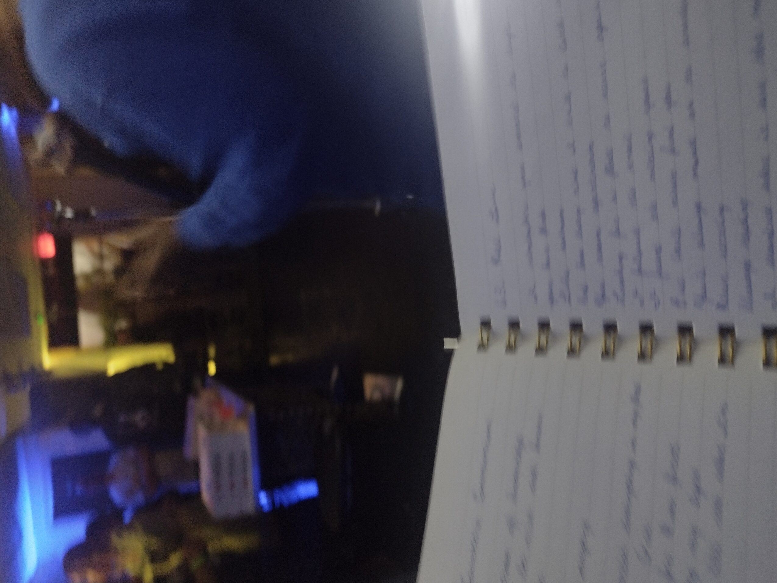 Open notebook with handwritten notes on a table, person in blue shirt in background, dim lighting.