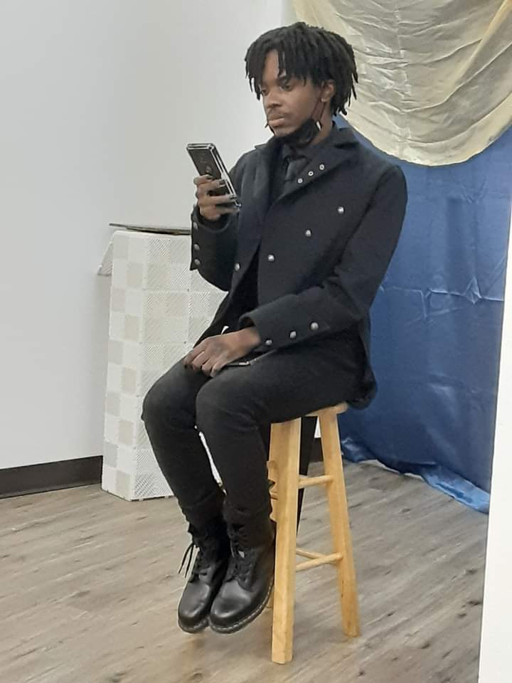 Person with dreadlocks sitting on a wooden stool, holding a phone, in a room with a blue curtain and a white wall.