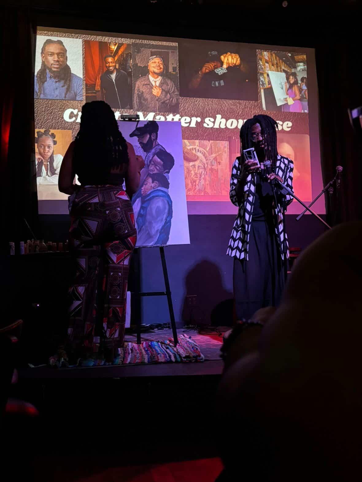 Two women stand on stage with a large screen behind them displaying images and text, in a dark room.