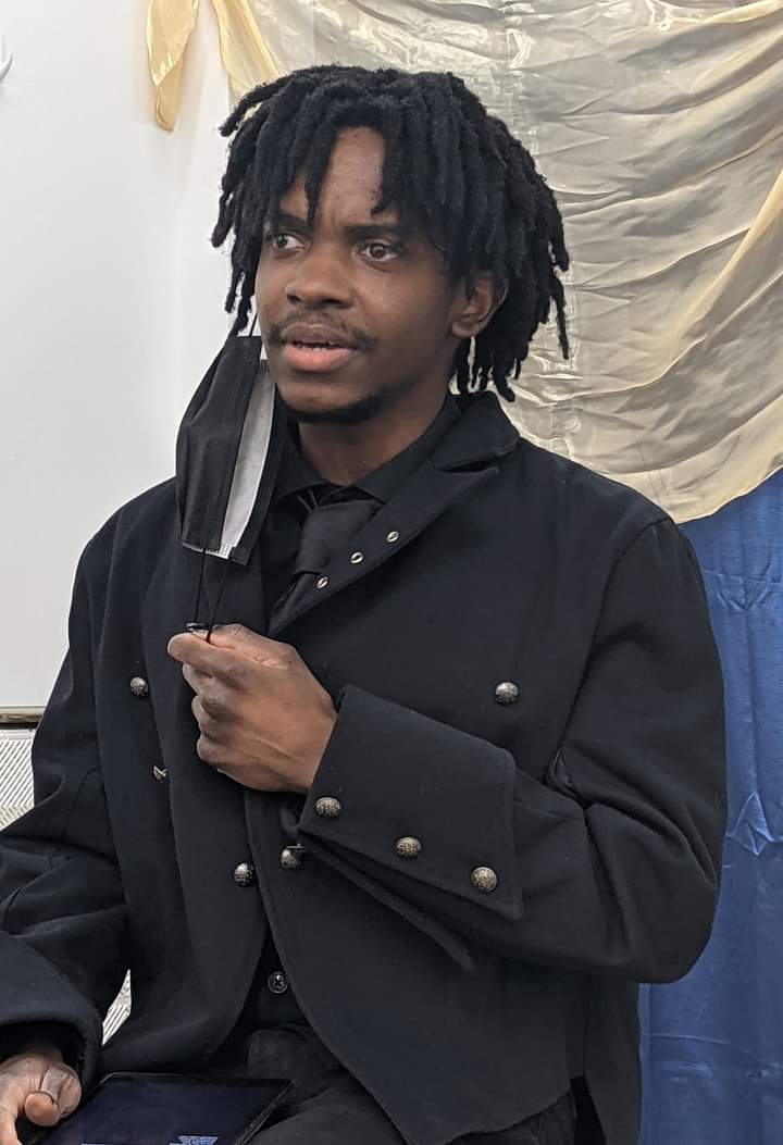 Young man with dreadlocks wearing a black jacket and holding a face mask, standing indoors with draped fabric in background.