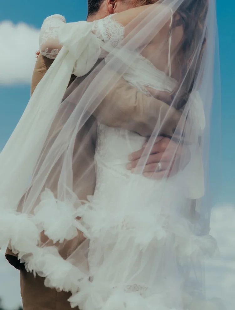 Couple embracing outdoors, woman in wedding dress with veil, sky background, romantic scene.