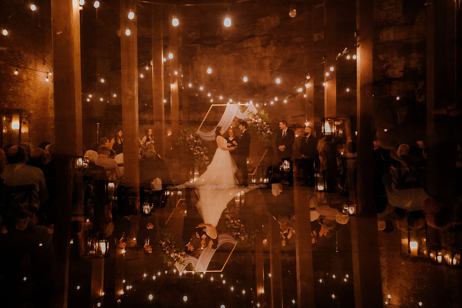 Wedding couple dancing in a warmly lit venue with guests and string lights, reflected on a shiny floor.