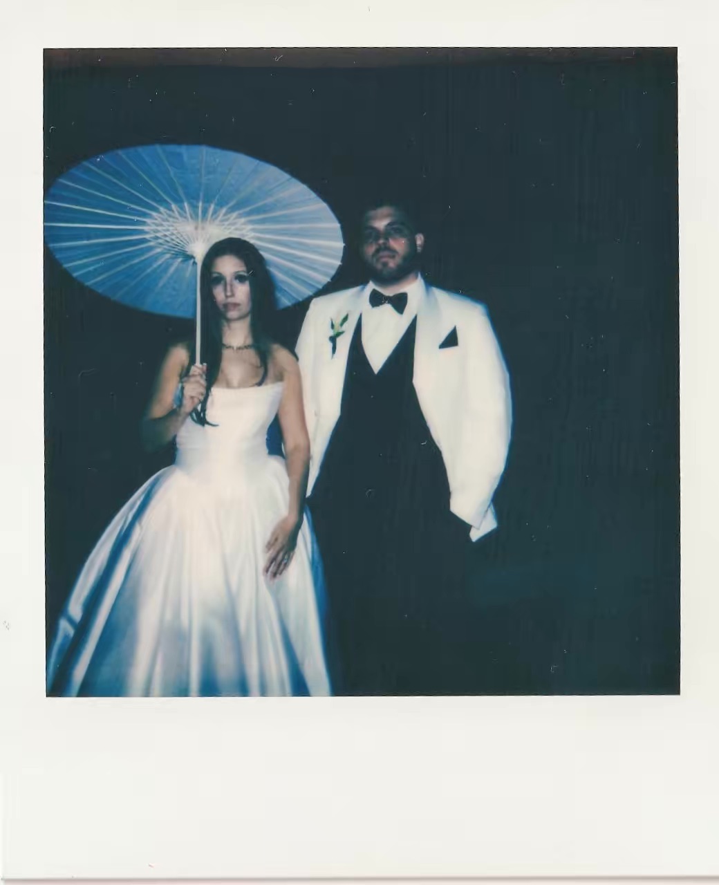 A man and woman in formal attire stand together, woman holding a parasol, against a dark background.
