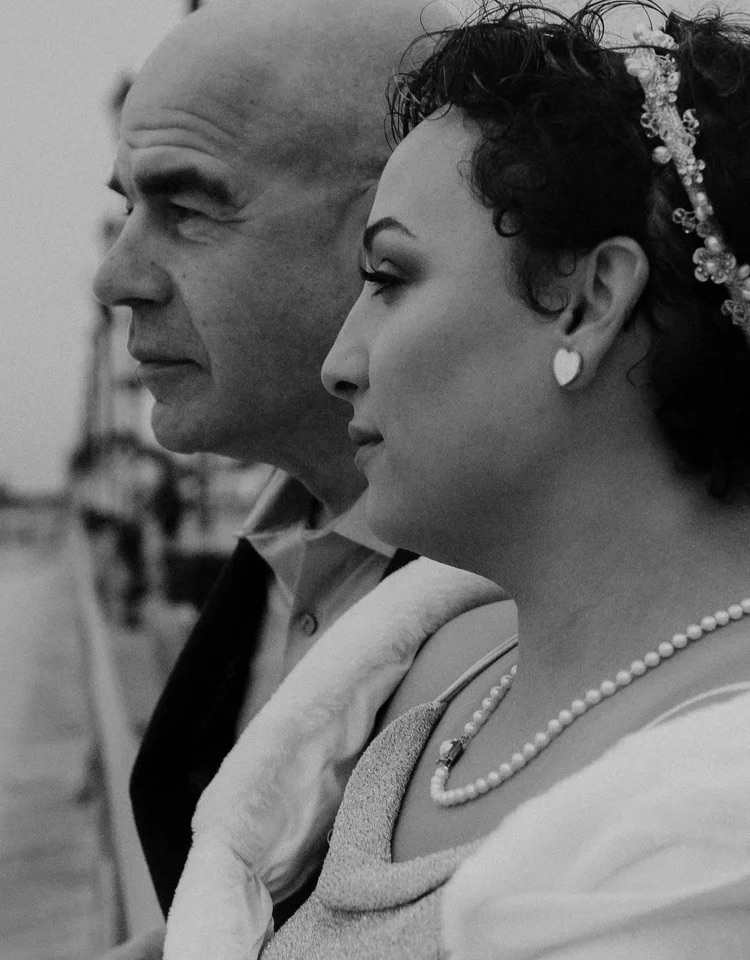 Two people, a man and a woman, are in profile view, looking to the right, with a blurred outdoor background.