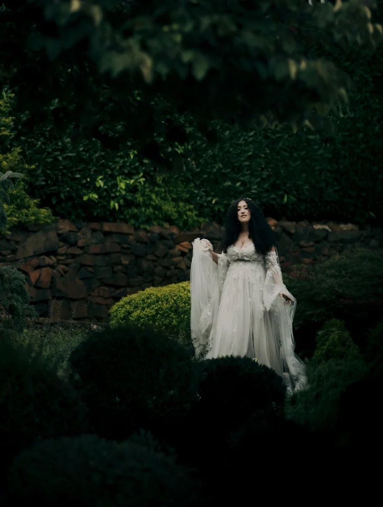 Woman in a flowing white dress standing outdoors among greenery and bushes, with a stone wall behind her.