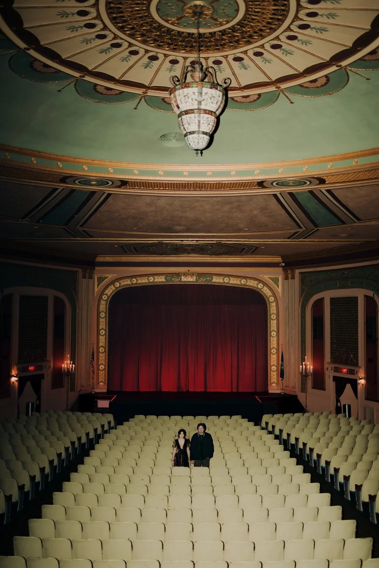 The theater has a red curtain, ornate architecture, and two people standing in the center of empty seats.