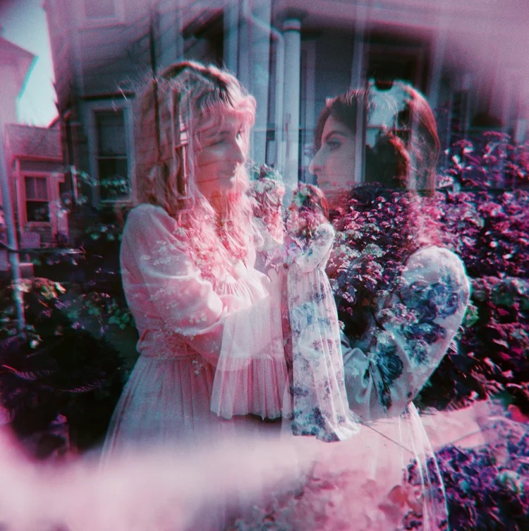 Two women face each other in a garden with flowers, appearing blurred and layered, with a pinkish hue.