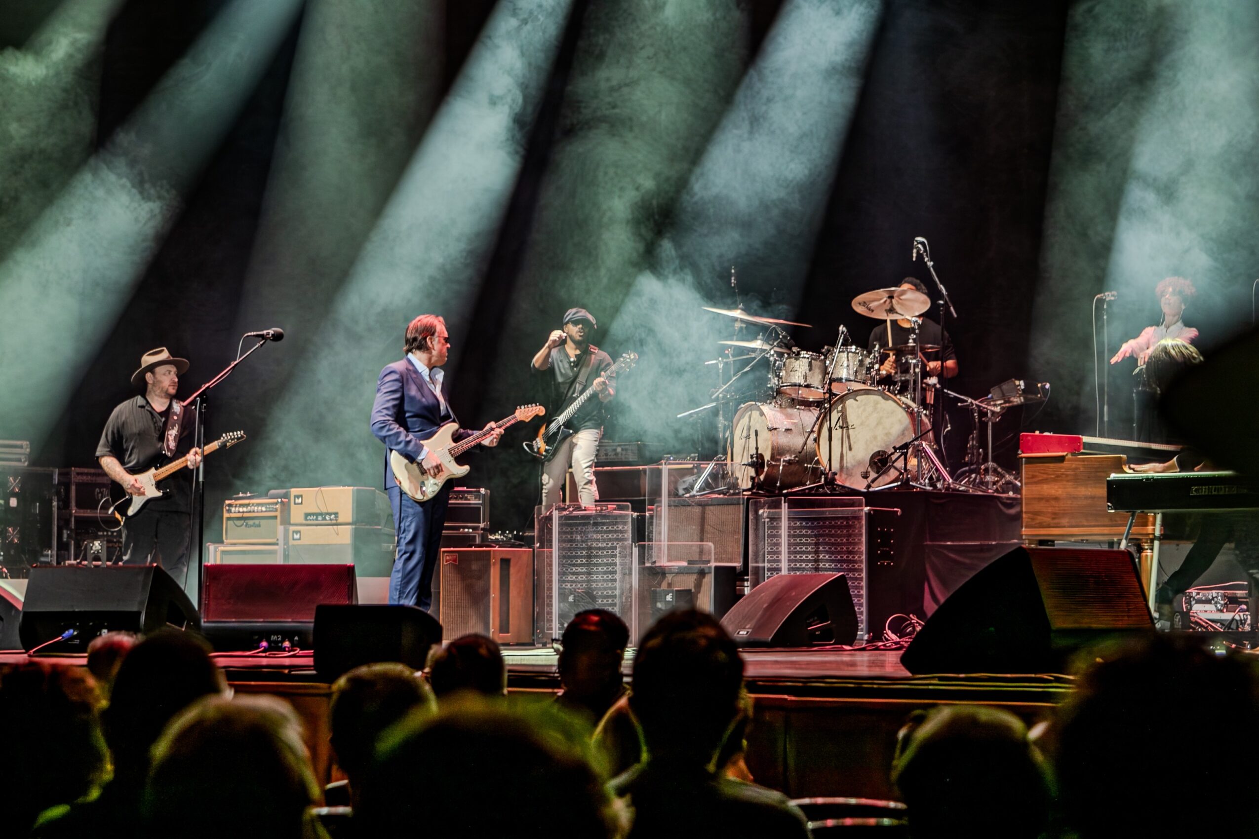 Band performs on stage with instruments, audience visible in foreground, stage lighting and smoke effects present.