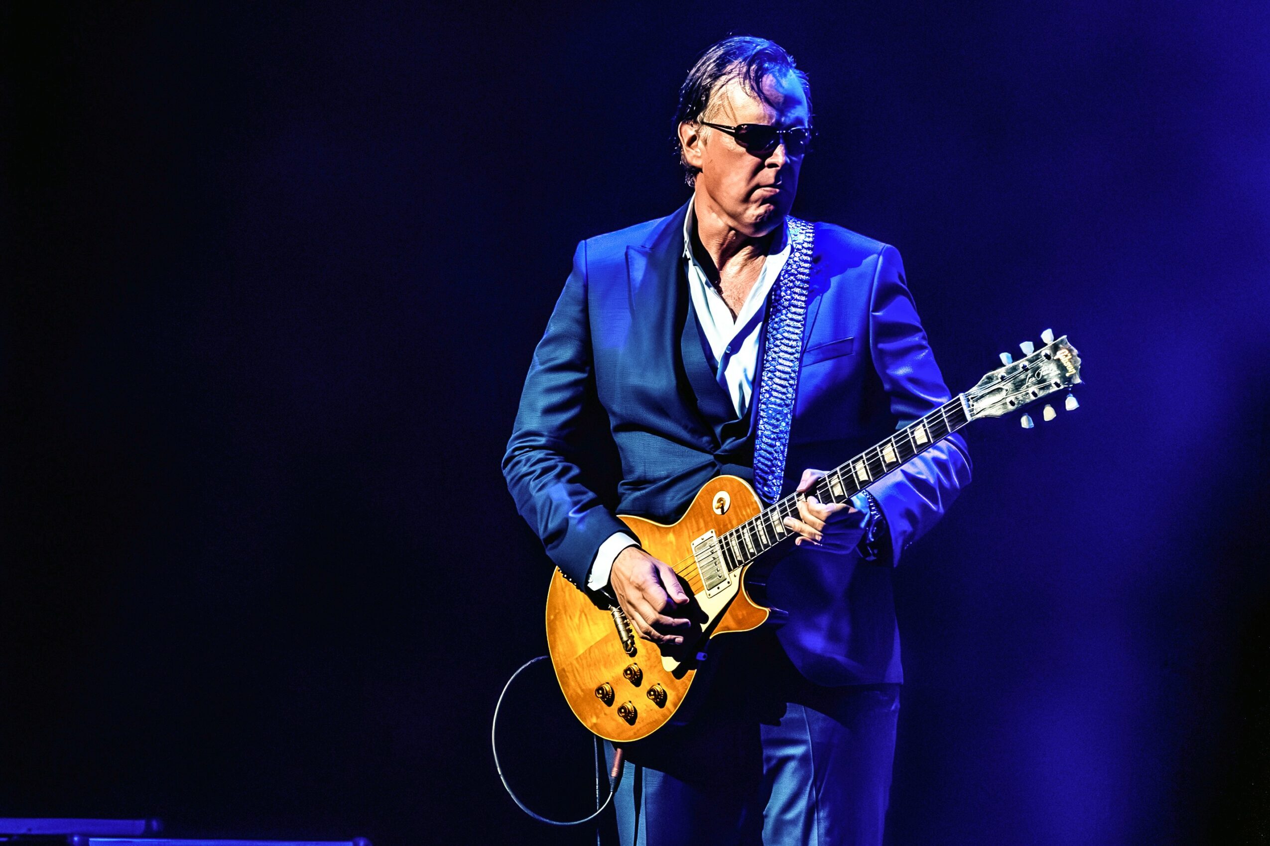 Man playing electric guitar on stage with dark background, wearing sunglasses and a blue suit.