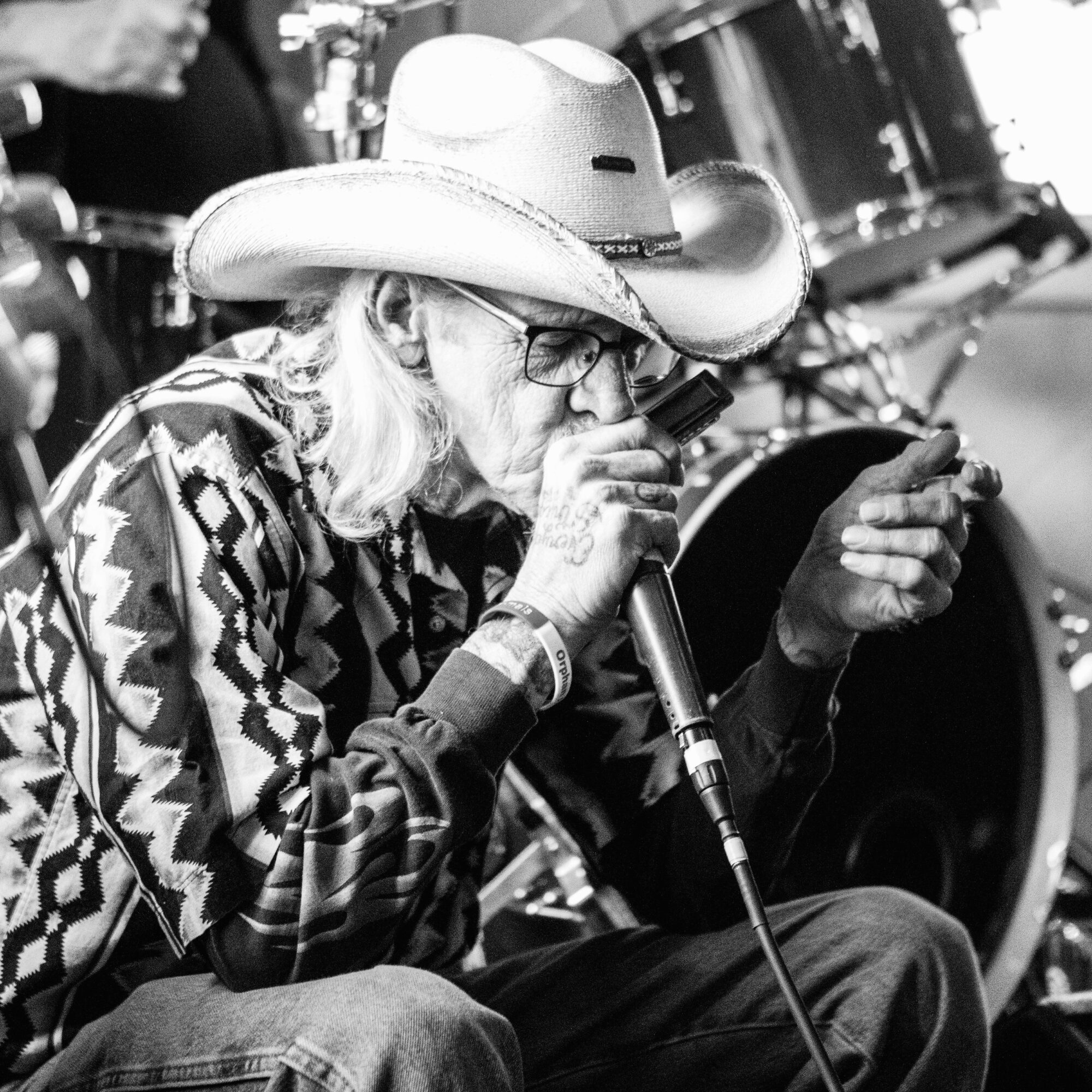Older man wearing a cowboy hat and glasses, singing into a microphone, with drums in the background.