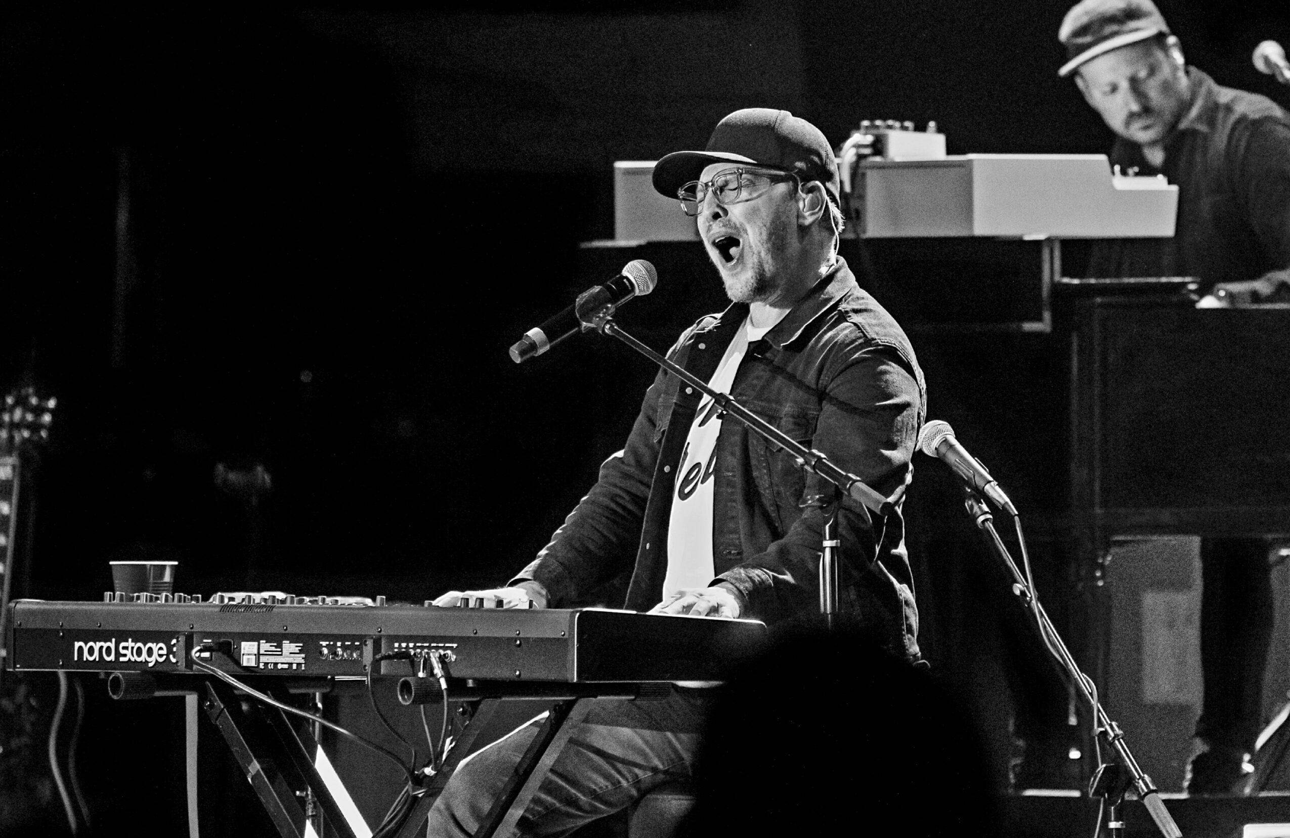 Musician singing and playing keyboard on stage with a microphone, with another person in the background, in black and white.