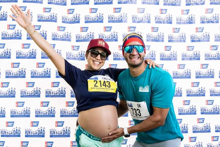 Pregnant woman and man smiling, posing with arms around each other, in front of a race backdrop, woman wearing a cap and sunglasses, man wearing sunglasses and a headband, both with race bibs.