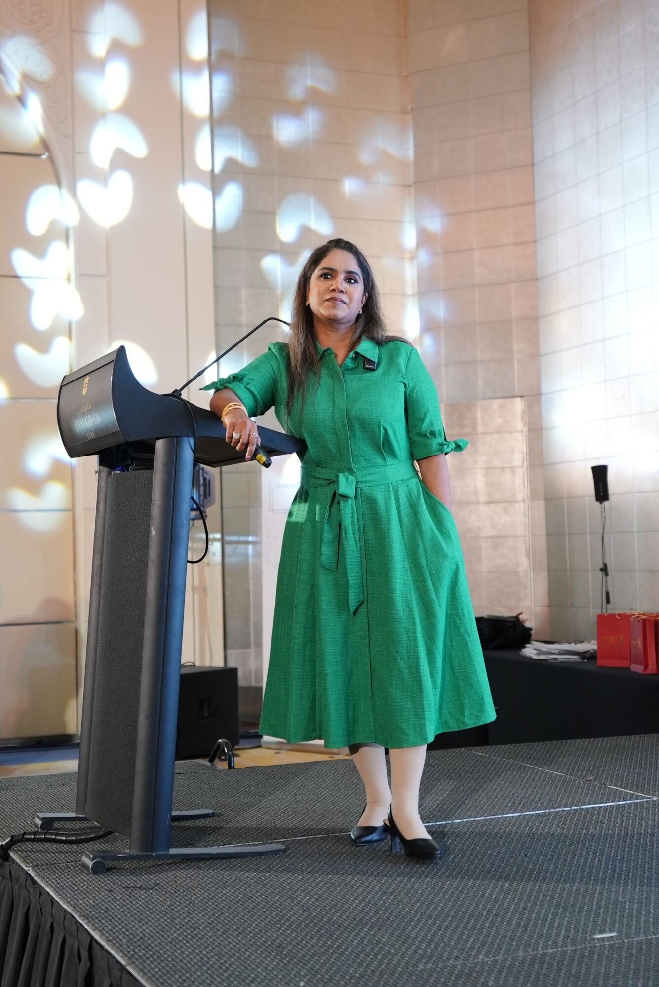 Woman in green dress standing on stage with a microphone, speaking to an audience, with stage lighting effects.