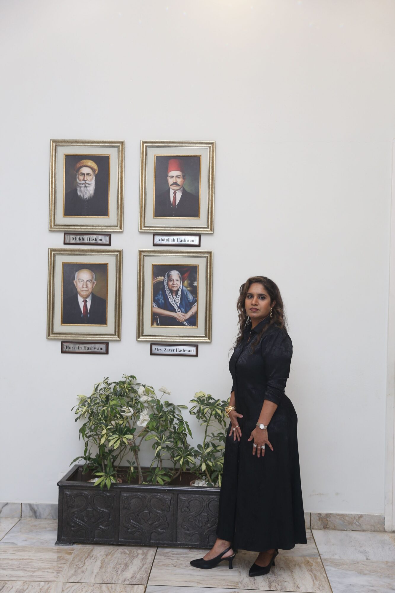 Woman in black dress standing in front of a white wall with four framed pictures, plants on a black stand, tiled floor