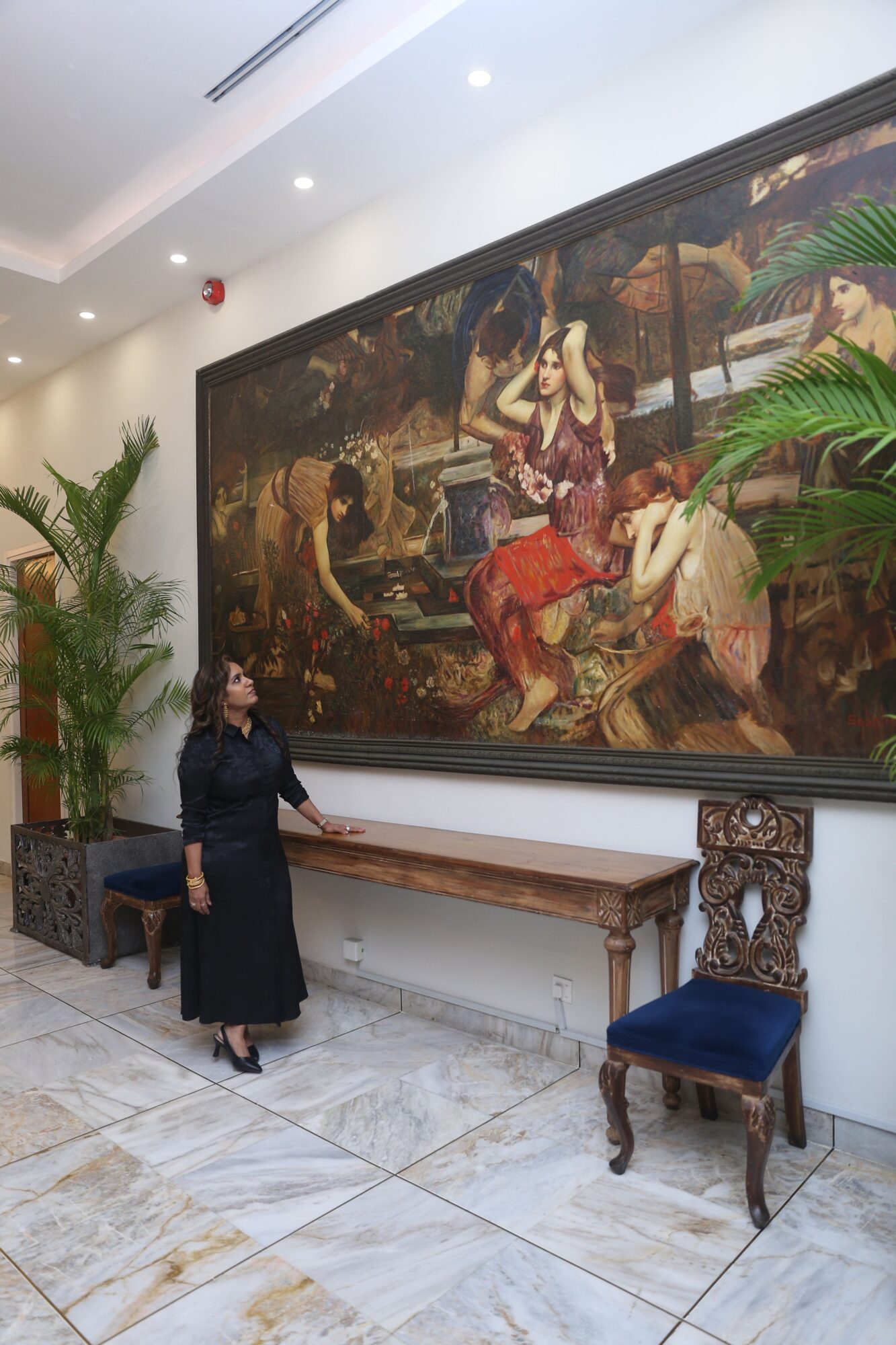 Person standing in a room with a large painting, a wooden bench, and a decorative chair, with plants and ceiling lights.