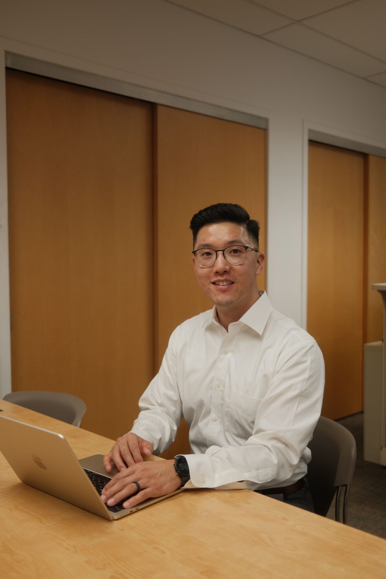 Man in white shirt sitting at a table with a laptop, hands on the keyboard, in a room with wooden doors.