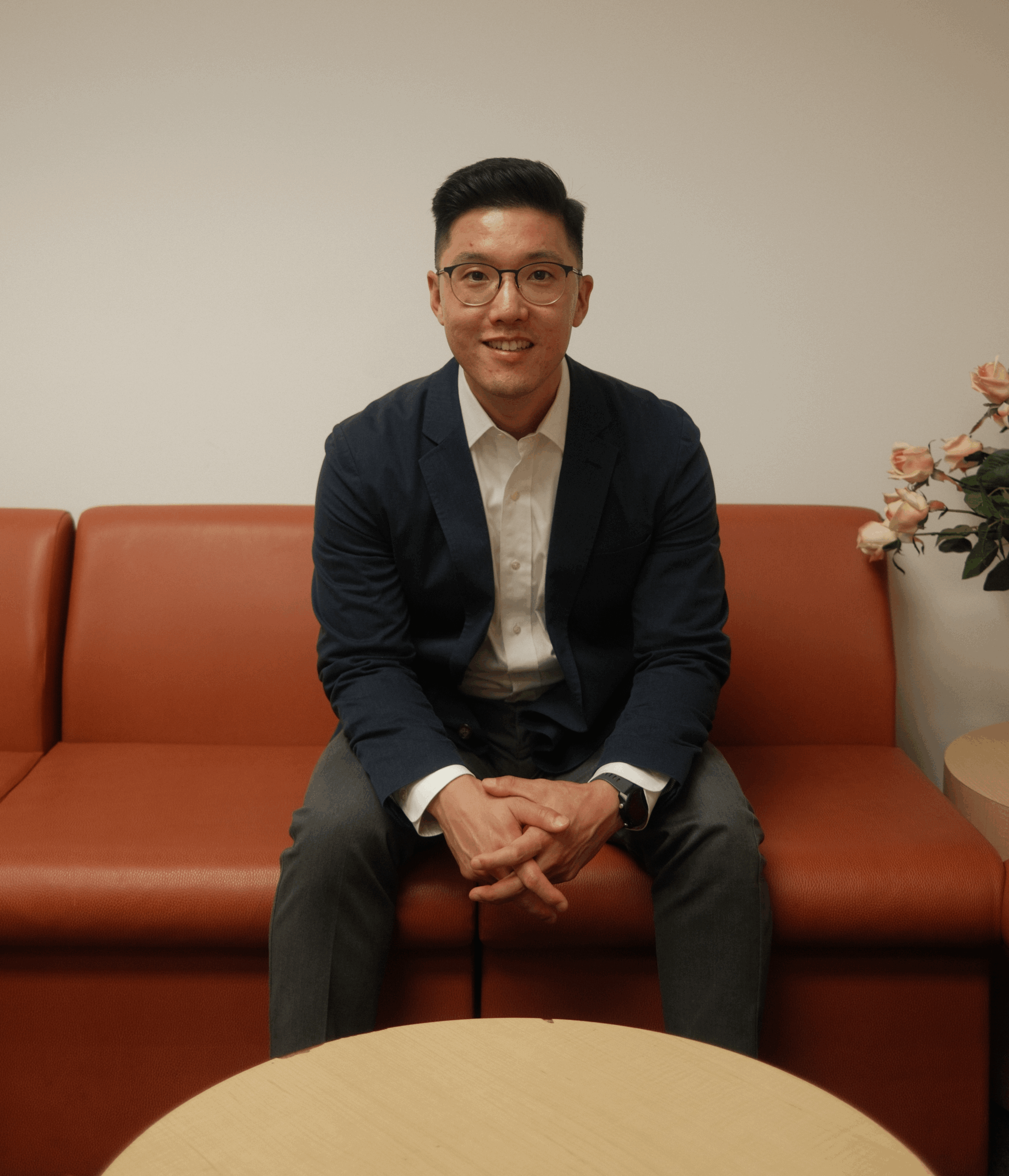 Man sitting on a brown sofa, smiling, wearing glasses, a dark blazer, and a white shirt, with a flower arrangement nearby.
