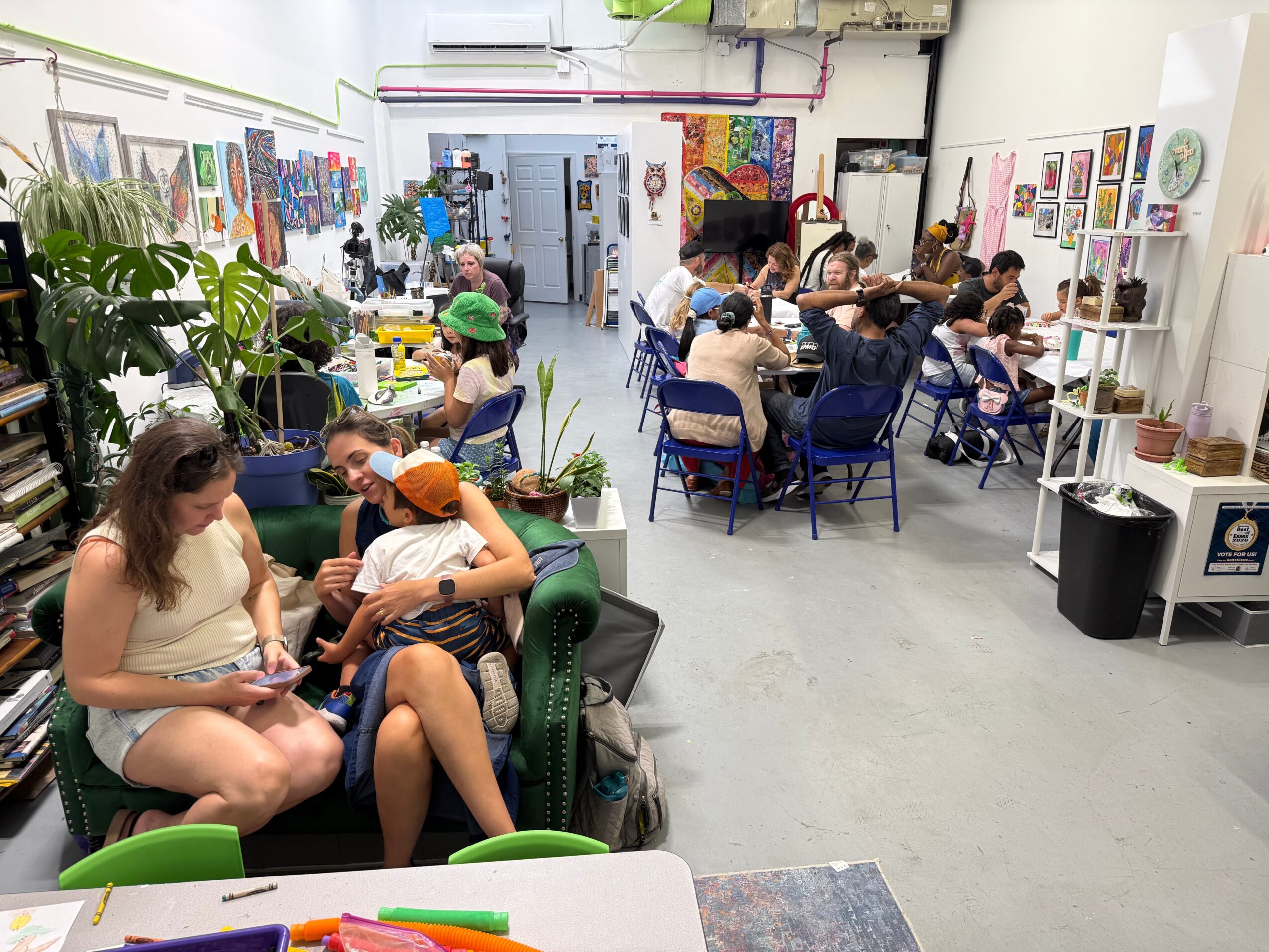 People sitting and socializing in an art gallery with colorful artwork on walls, plants, and tables, some using laptops or phones.