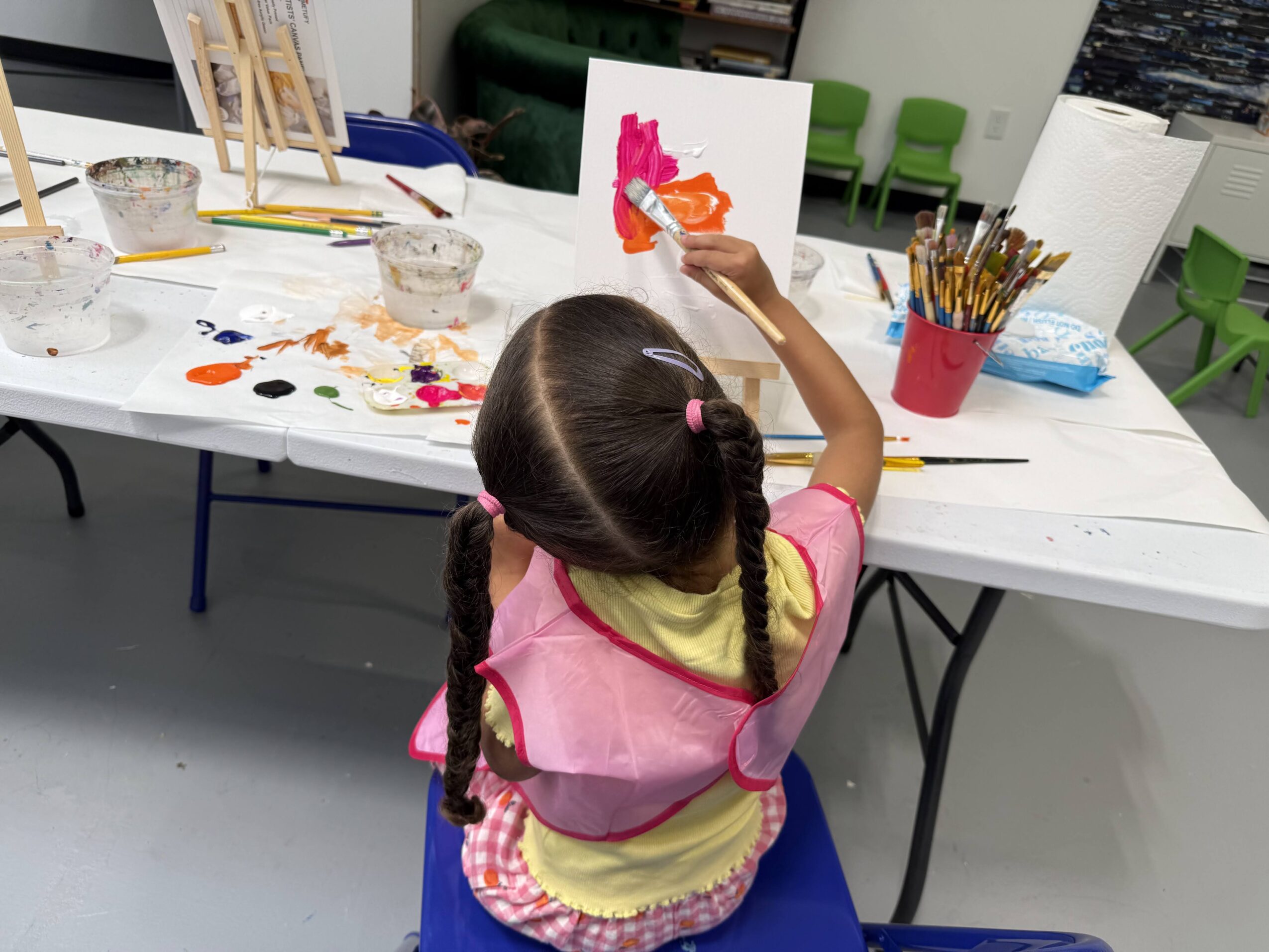 Child with braided hair painting on canvas at art table, surrounded by art supplies.