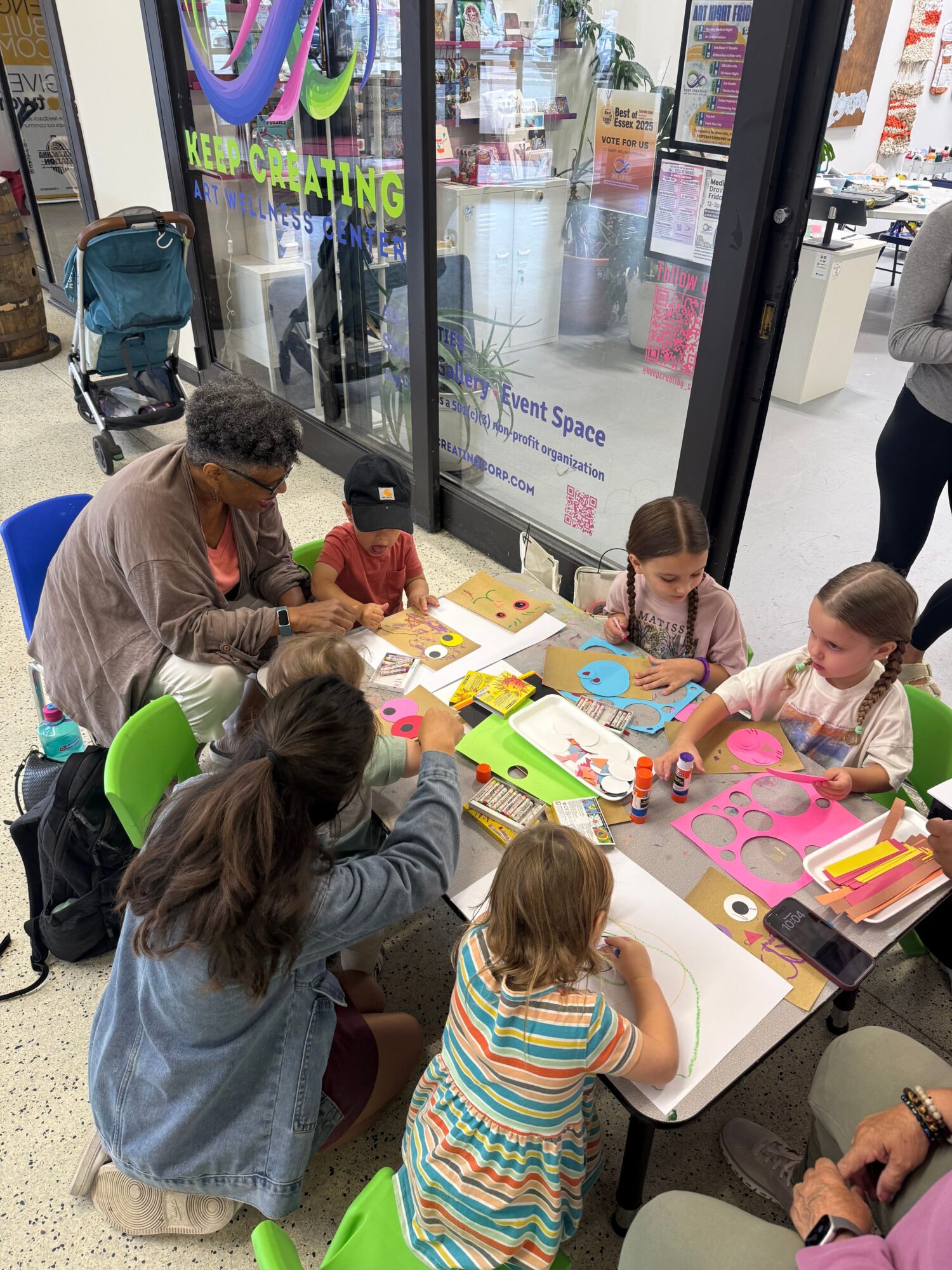 Children and adults sit around a table with colorful craft supplies, working on arts and crafts projects indoors.