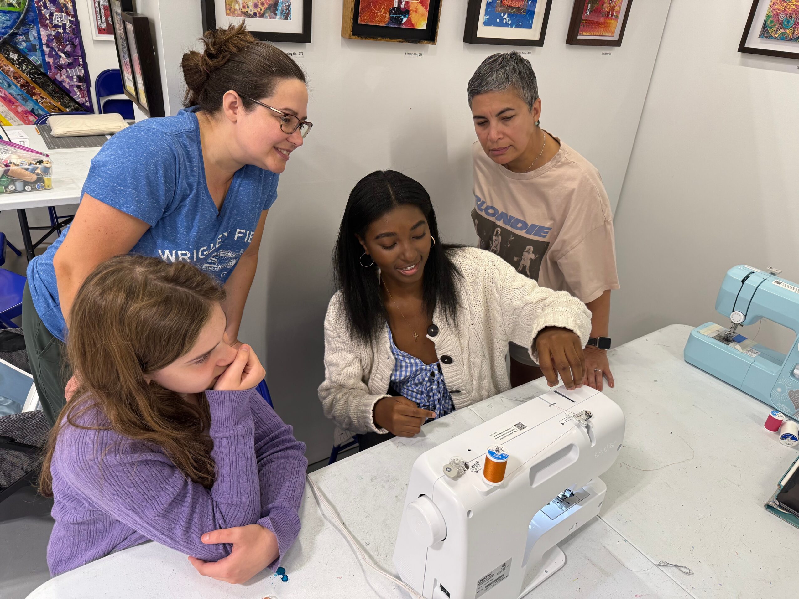 Four people gathered around a sewing machine, with framed pictures on the wall behind them.