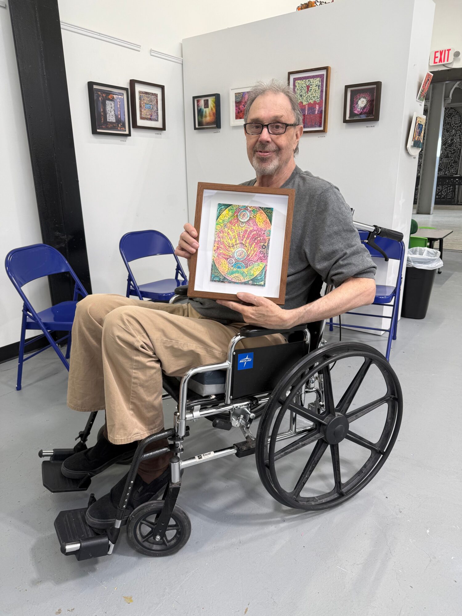 Person in a wheelchair holding a colorful framed artwork, in a room with framed pictures on the wall.