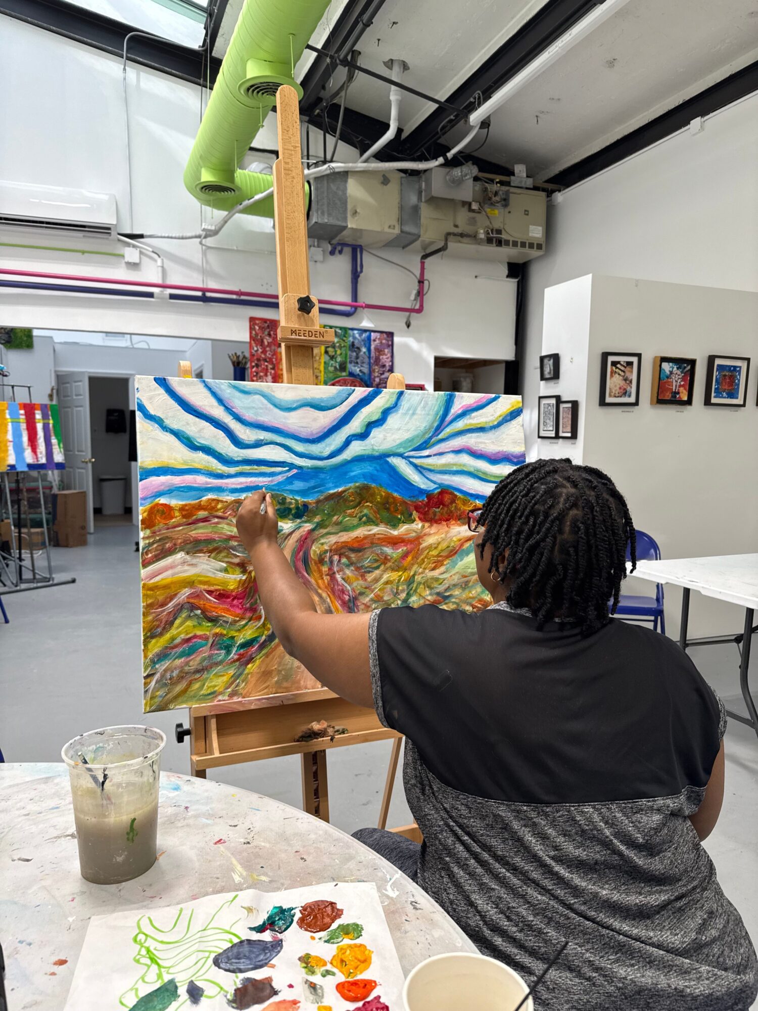Person with dreadlocks painting colorful abstract artwork on canvas in studio, with art supplies and a glass of water nearby.