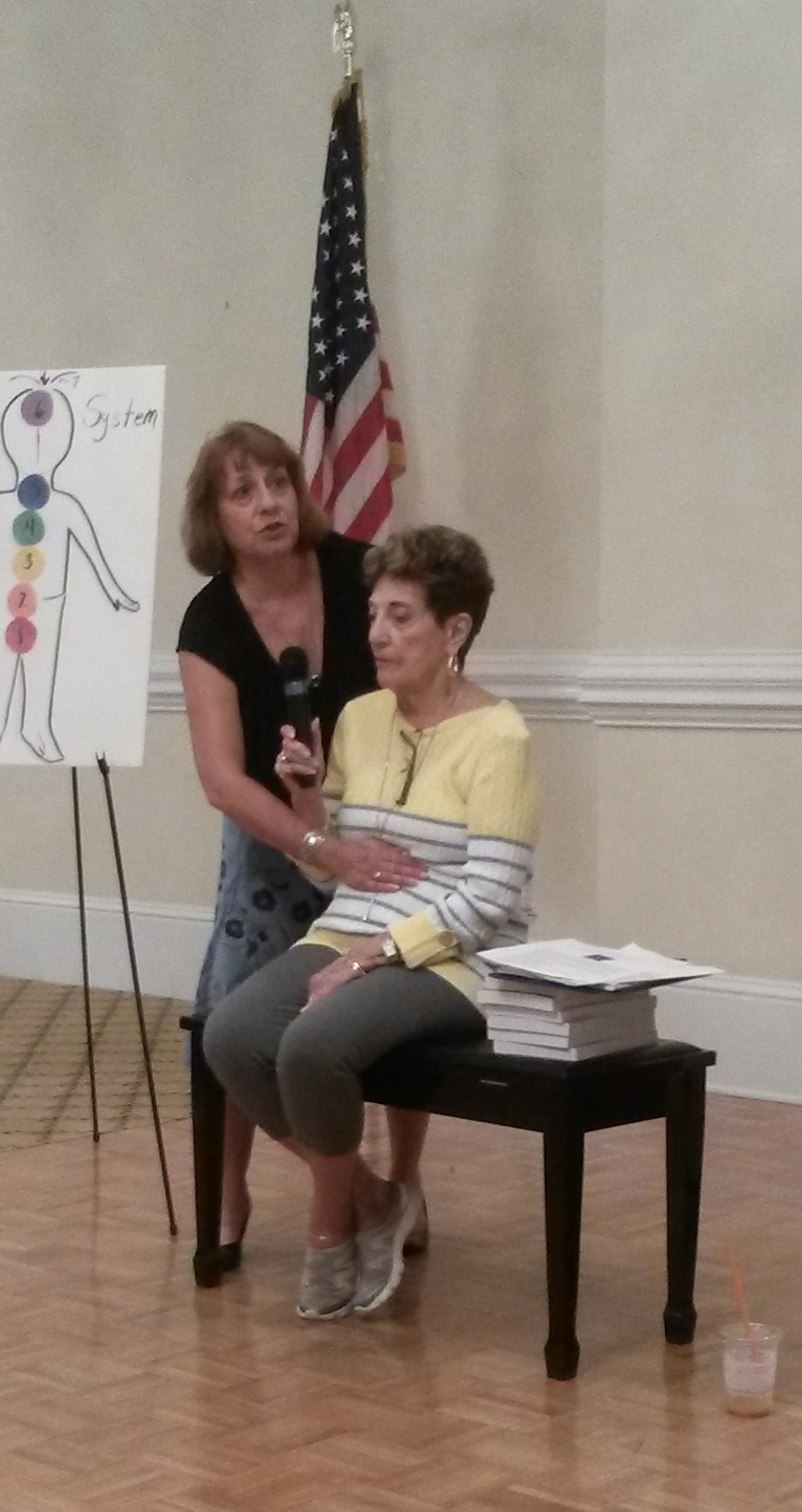 Two women in a room with an American flag, one sitting on a chair and the other standing beside her, with a chart on a stand.