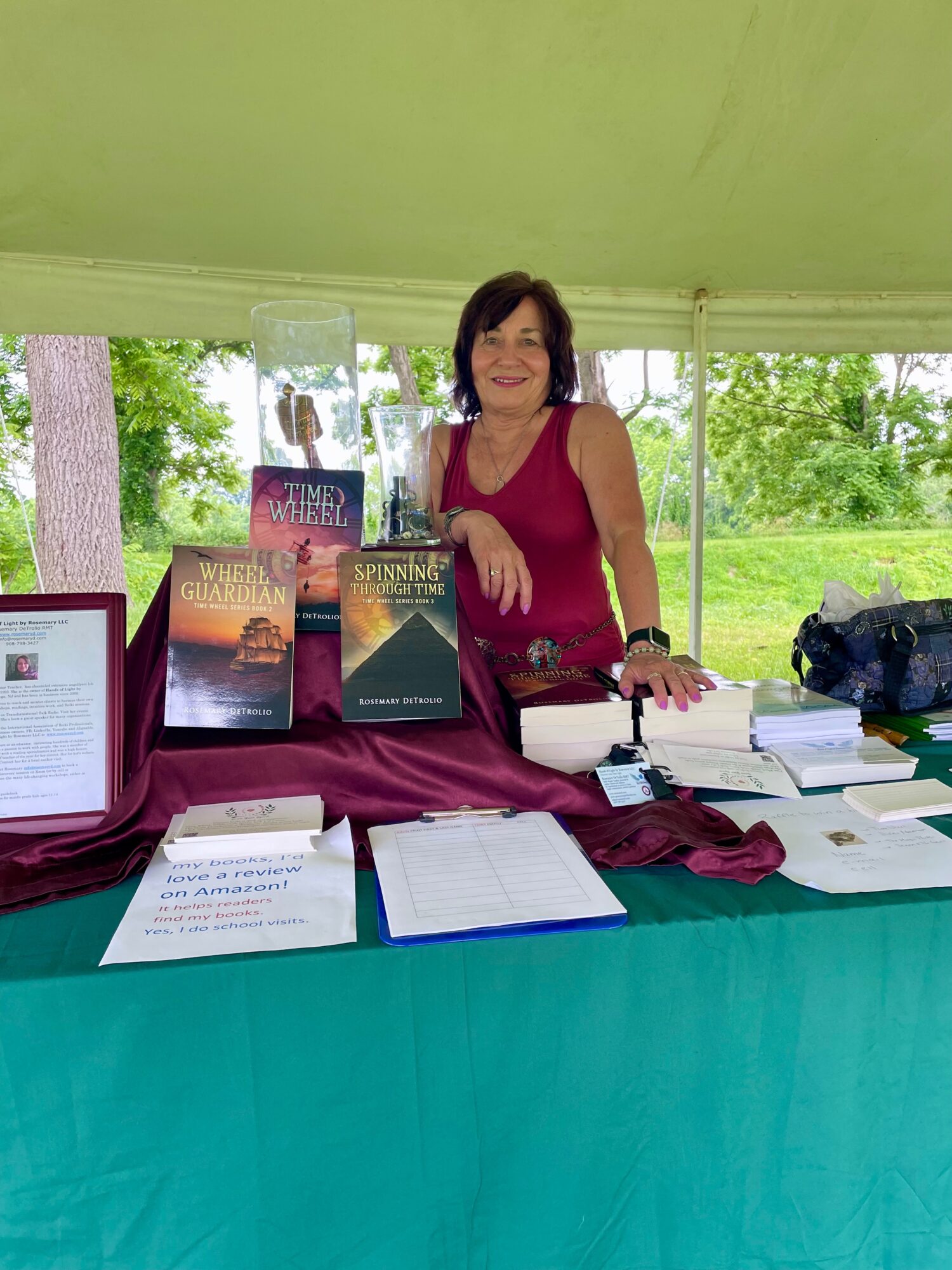 Woman standing behind a table with books and papers, outdoors with trees and greenery in background.