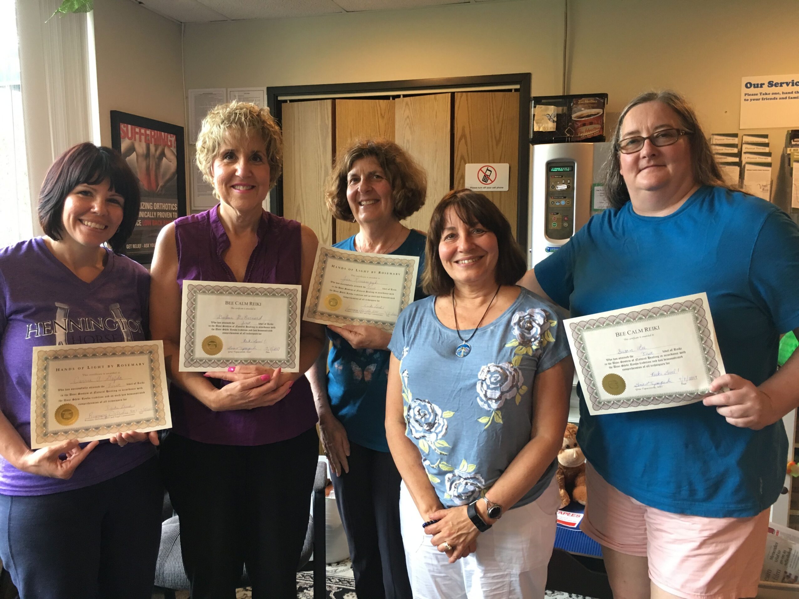 Group of five people holding certificates, standing indoors, smiling, with a window and framed pictures in the background.