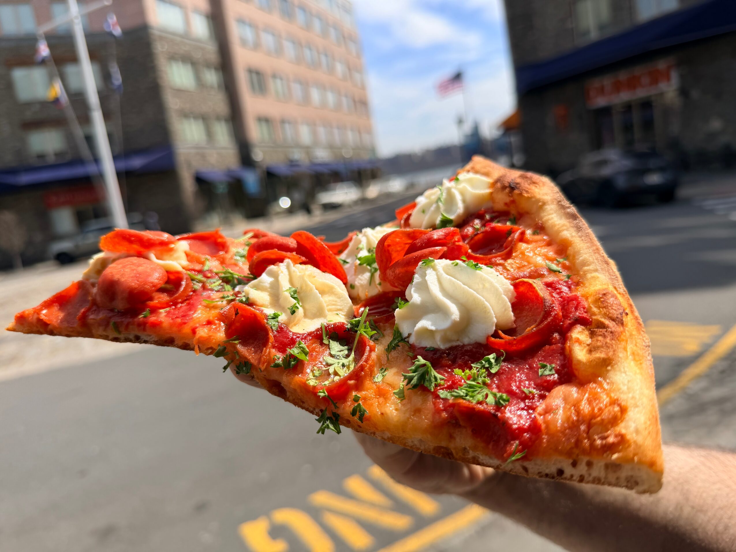 A slice of pizza with toppings including cheese, tomatoes, and herbs held in a person's hand outdoors.