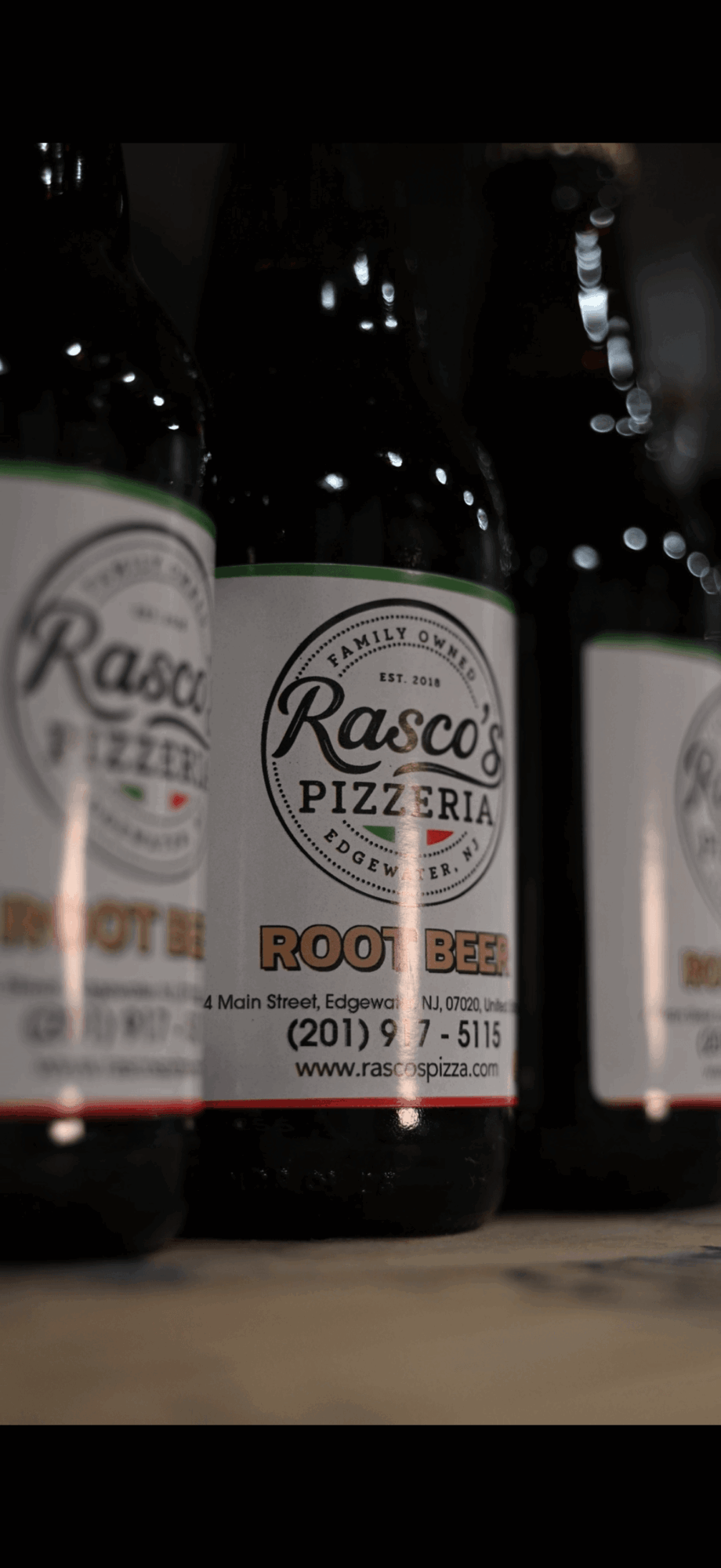 Three bottles of Rasco's Pizzeria root beer on a shelf, with one bottle in focus and others blurred in background.