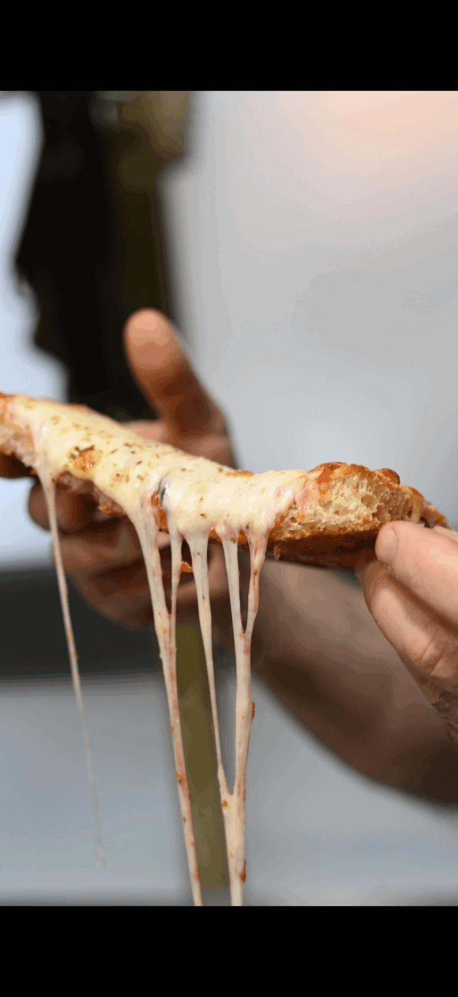 Person holding a slice of cheesy pizza with melted cheese stretching, against a blurred background.