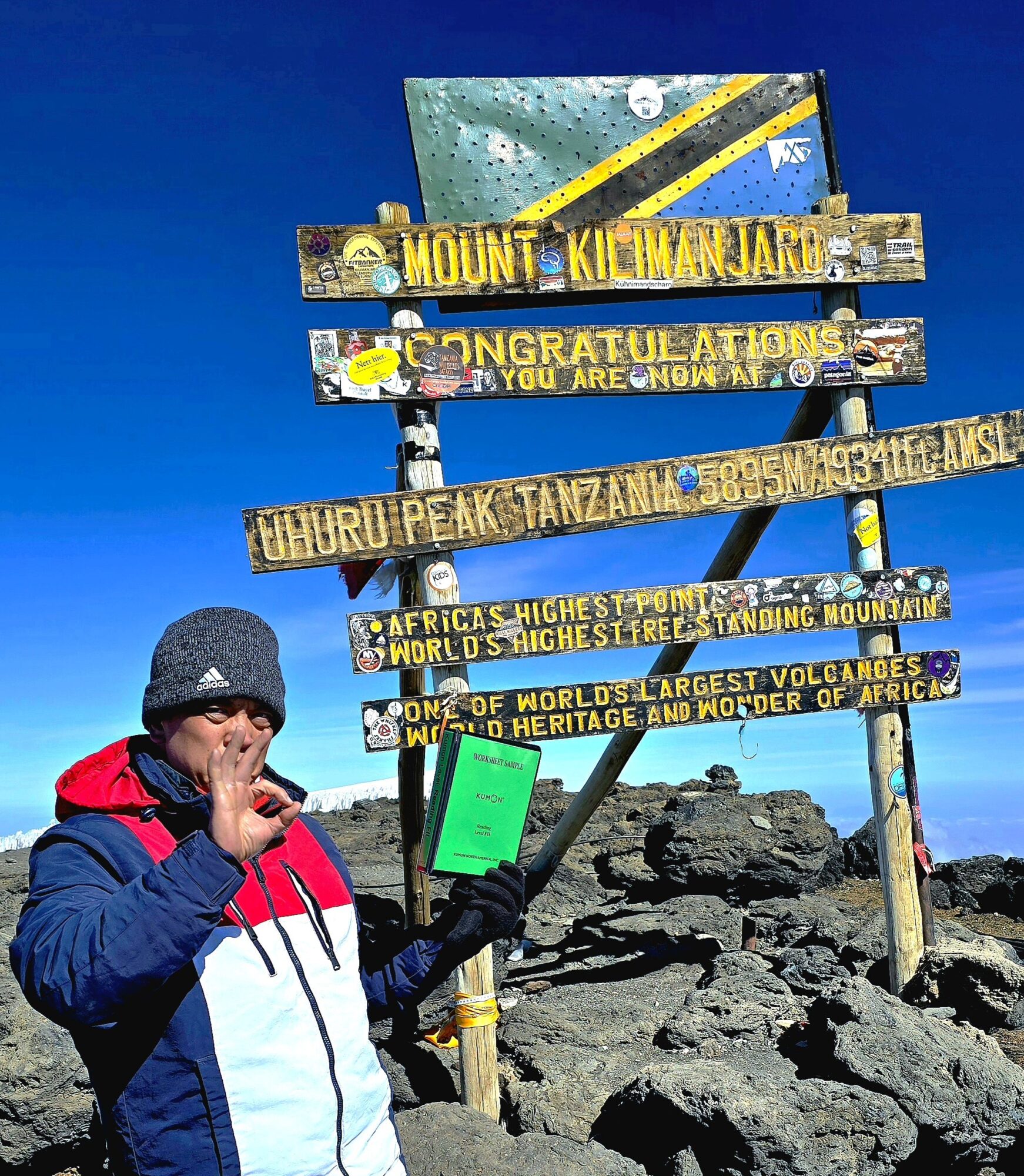 Person in winter clothing holding a green sign at Mount Kilimanjaro summit with a blue sky background.
