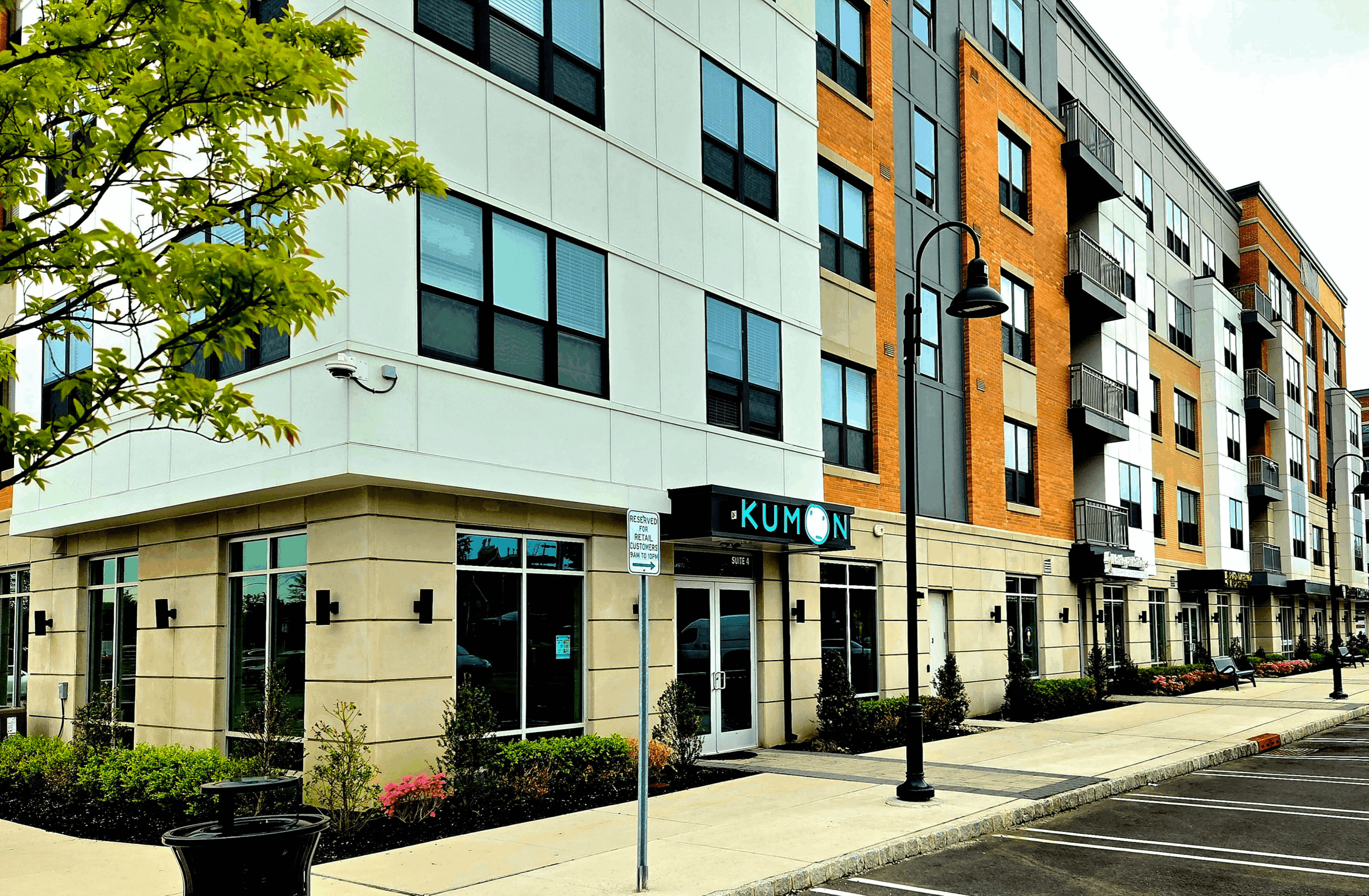 Modern multi-story building with large windows and a Kumon learning center on the ground floor, sidewalk, street, and trees.