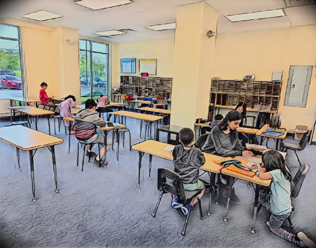 Children sitting at tables in a classroom with large windows, bookshelves, and a teacher assisting students.