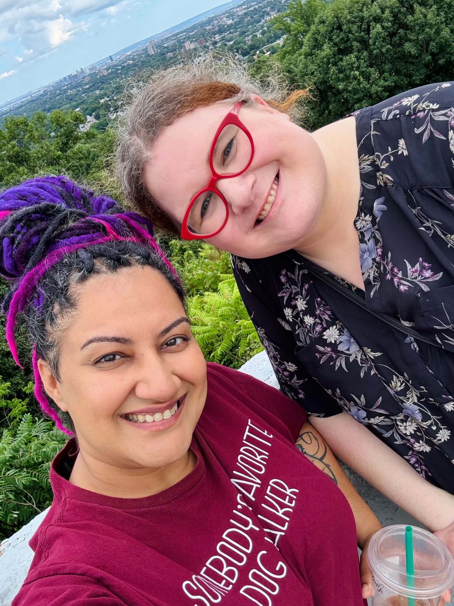 Two women smiling outdoors with greenery and a cityscape in the background, one with purple dreadlocks and the other with red glasses.