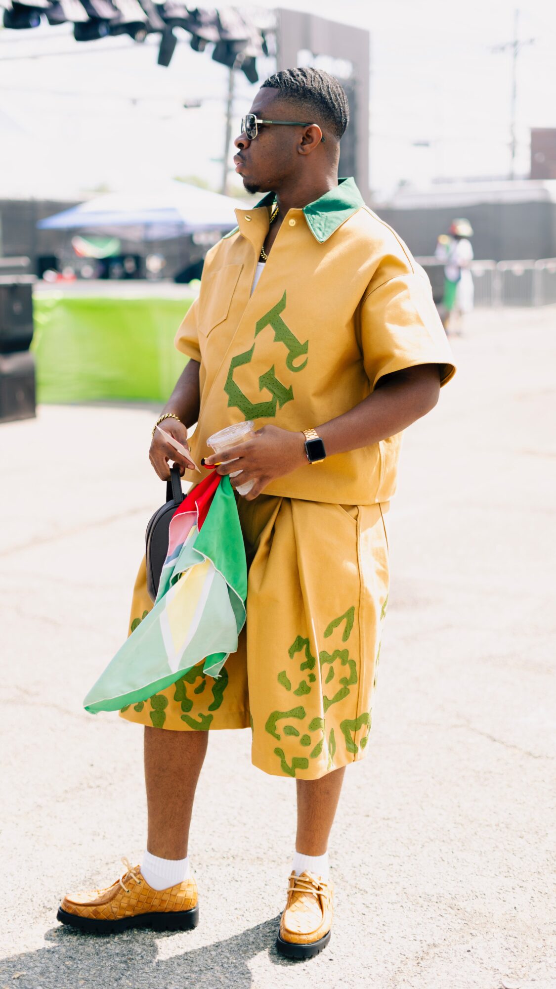 Person standing outdoors holding a flag, wearing yellow outfit with green patterns, sunglasses, and sneakers.