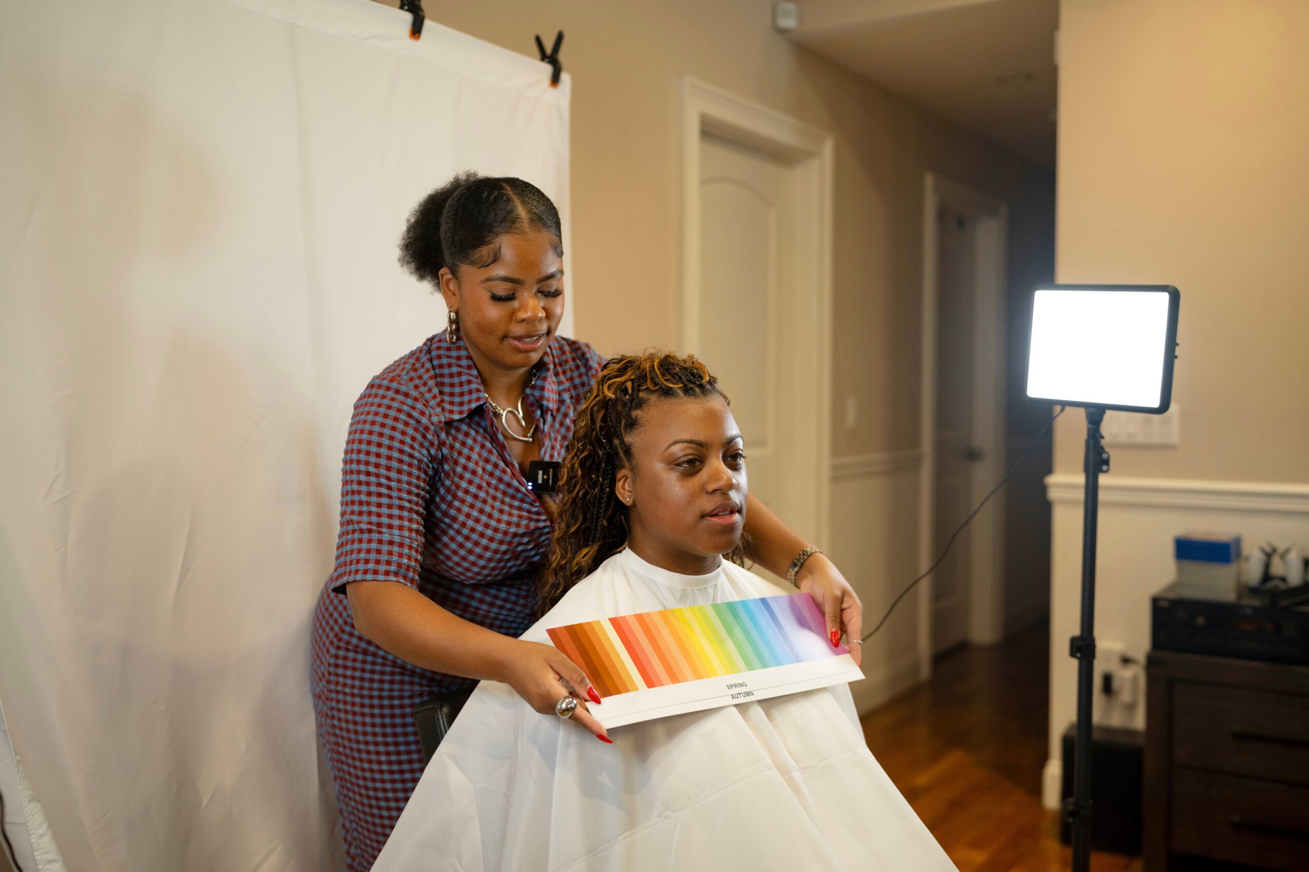 Two women, one seated and one standing, in a room with a white backdrop, holding a color palette. Bright light nearby.