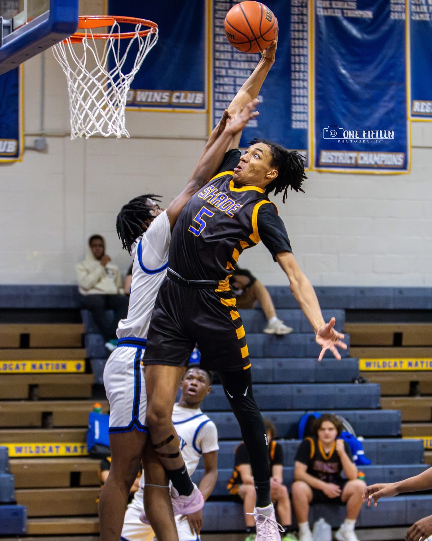 Two basketball players compete for the ball near the hoop, with spectators seated in the background.