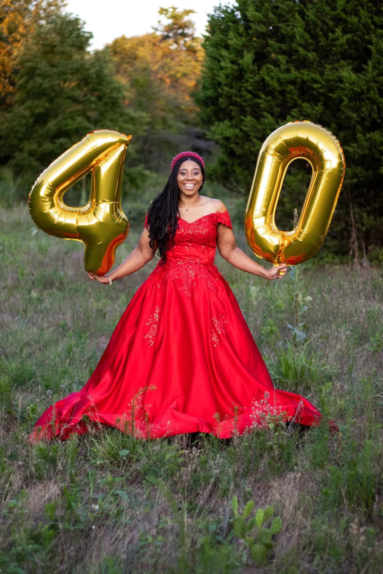 Woman in red dress holding gold balloons shaped as 4 and 0 in a grassy outdoor setting.