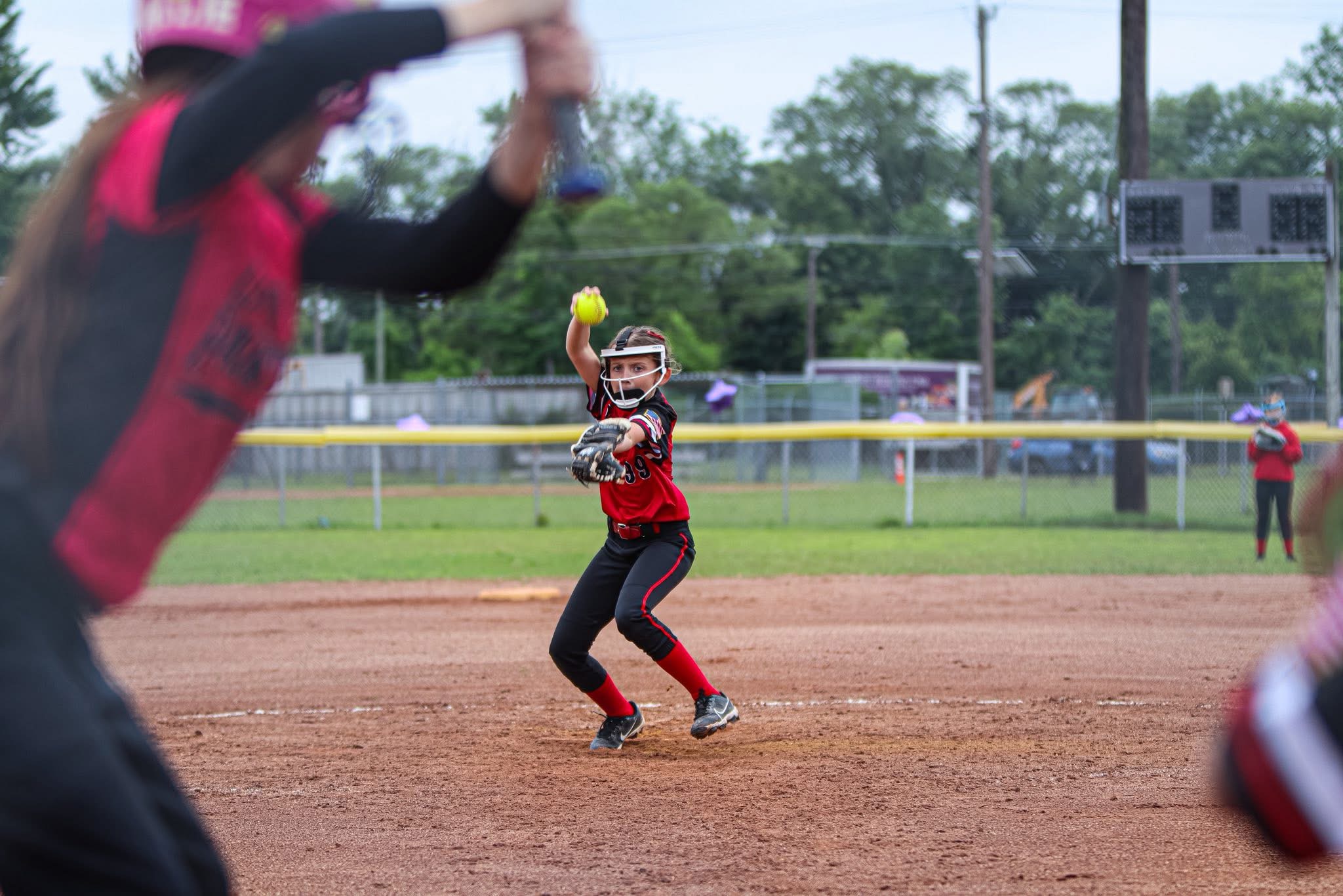 Child in red and black uniform catching a yellow ball during a softball game on a dirt field.