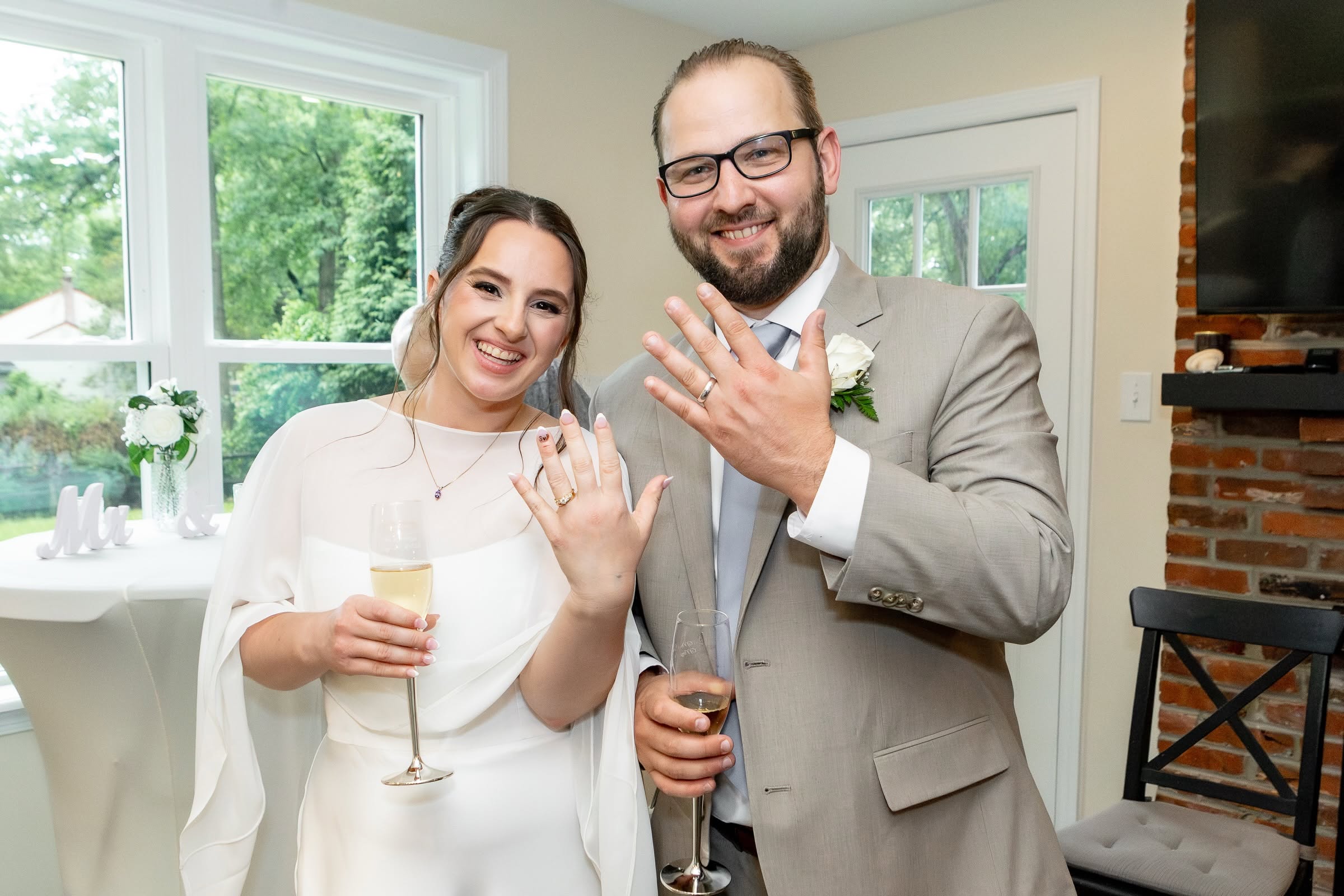 A smiling couple shows off their wedding rings indoors, holding drinks, with a window and fireplace in the background.