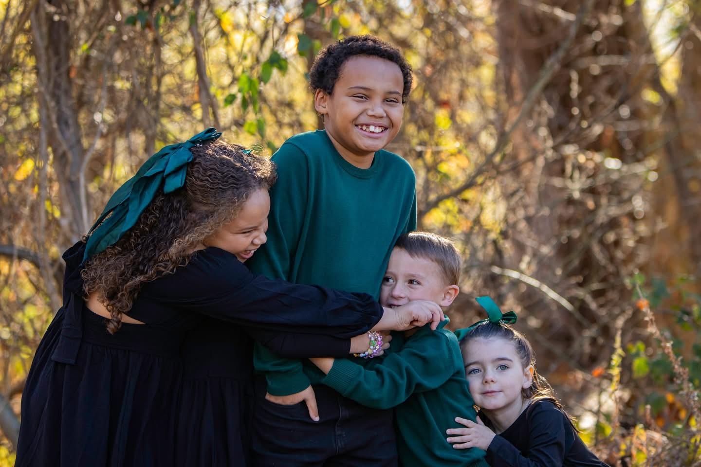 A woman and three children outdoors, smiling and playing, surrounded by trees and autumn foliage.