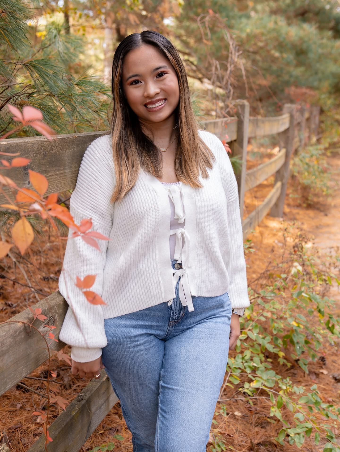 Young woman with long hair smiling outdoors, standing near a wooden fence with autumn foliage.